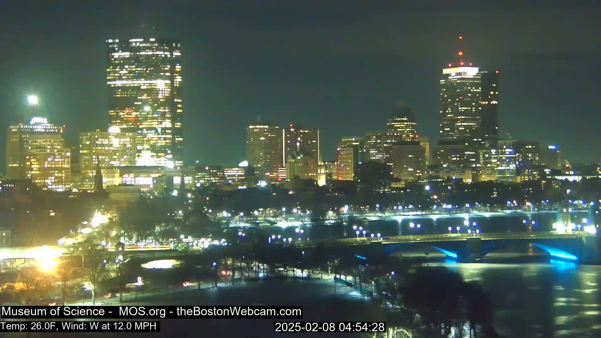 A nighttime cityscape with brightly lit buildings across a river, under a clear sky with a 26°F temperature and a 12 mph west wind.