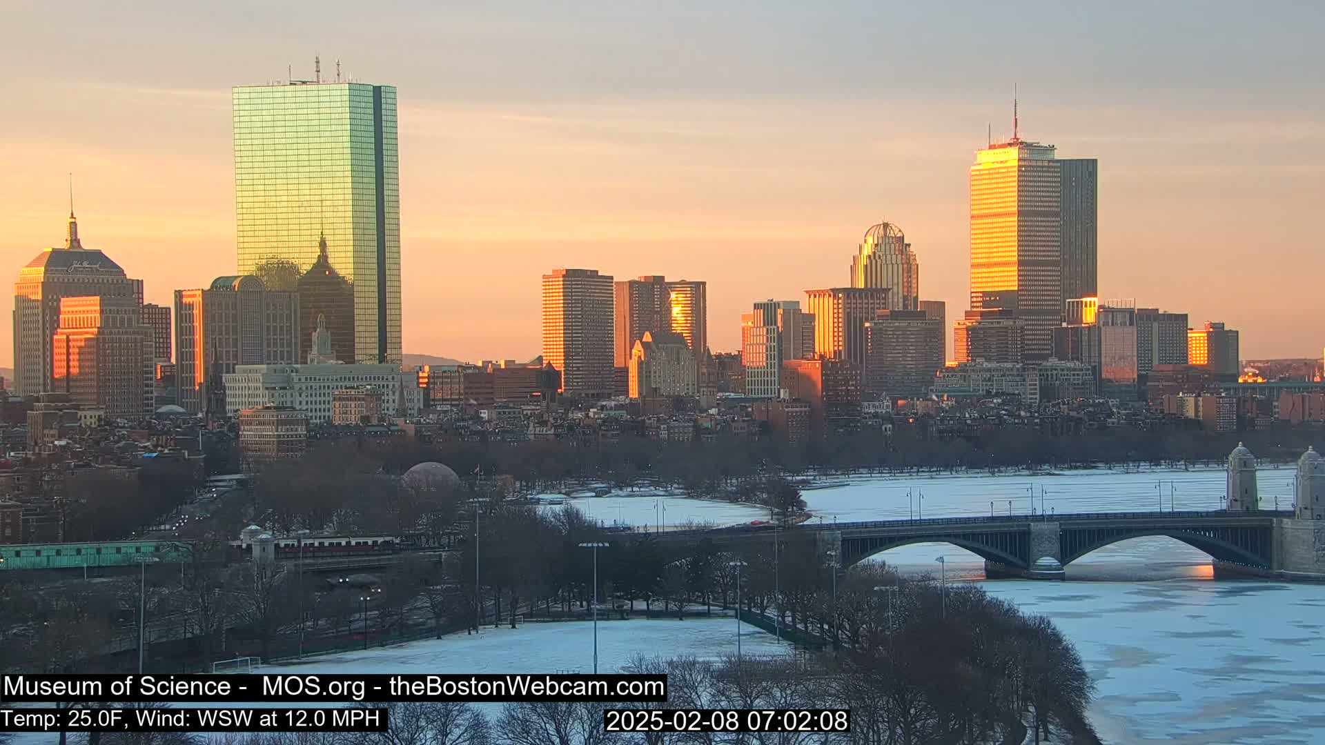 A city skyline at sunrise, with a partially frozen river and bridge in the foreground, under a clear, cold sky.