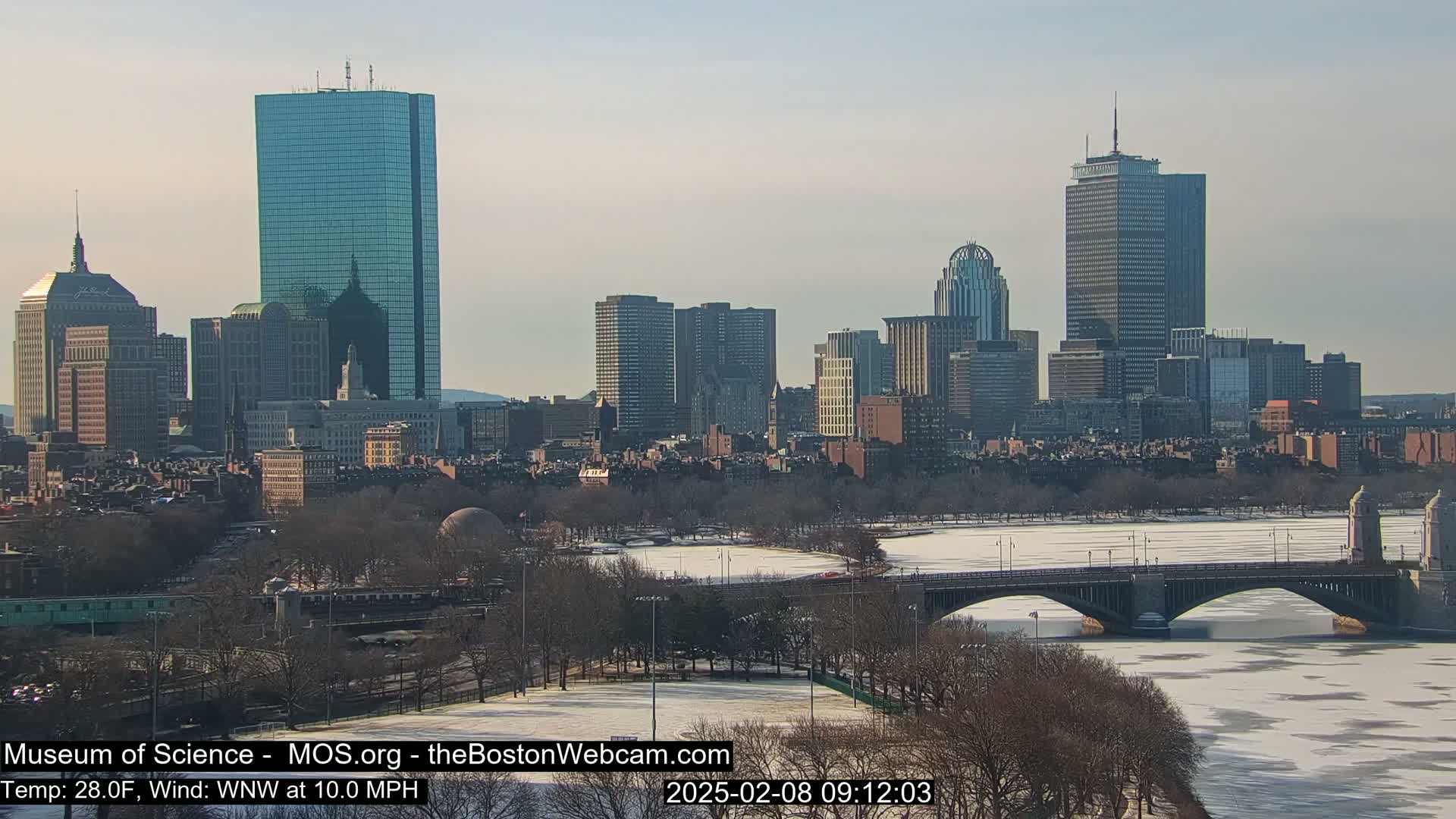 A city skyline with several skyscrapers is viewed from across a partially frozen river, under a clear, sunny sky.