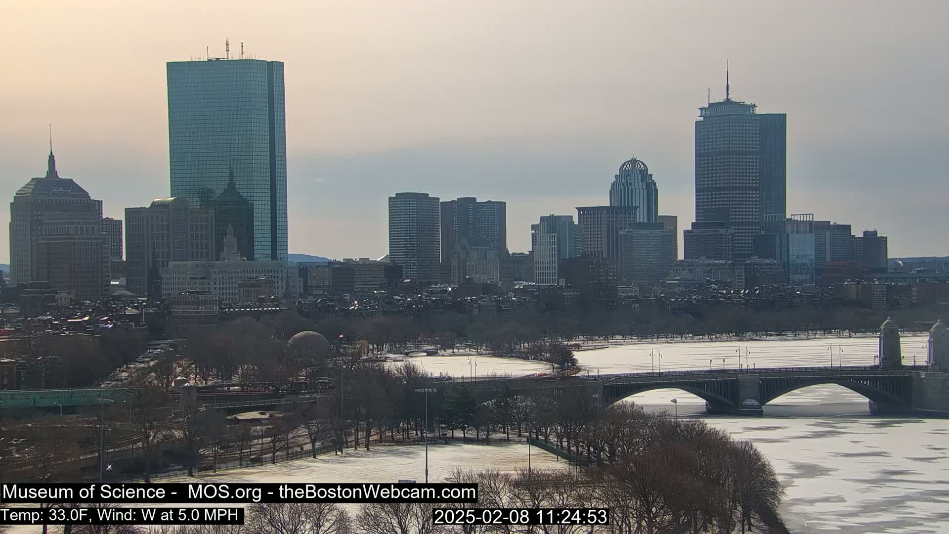 A snow-covered Boston cityscape, including several skyscrapers and a bridge over a partially frozen river, is viewed on a partly cloudy day.