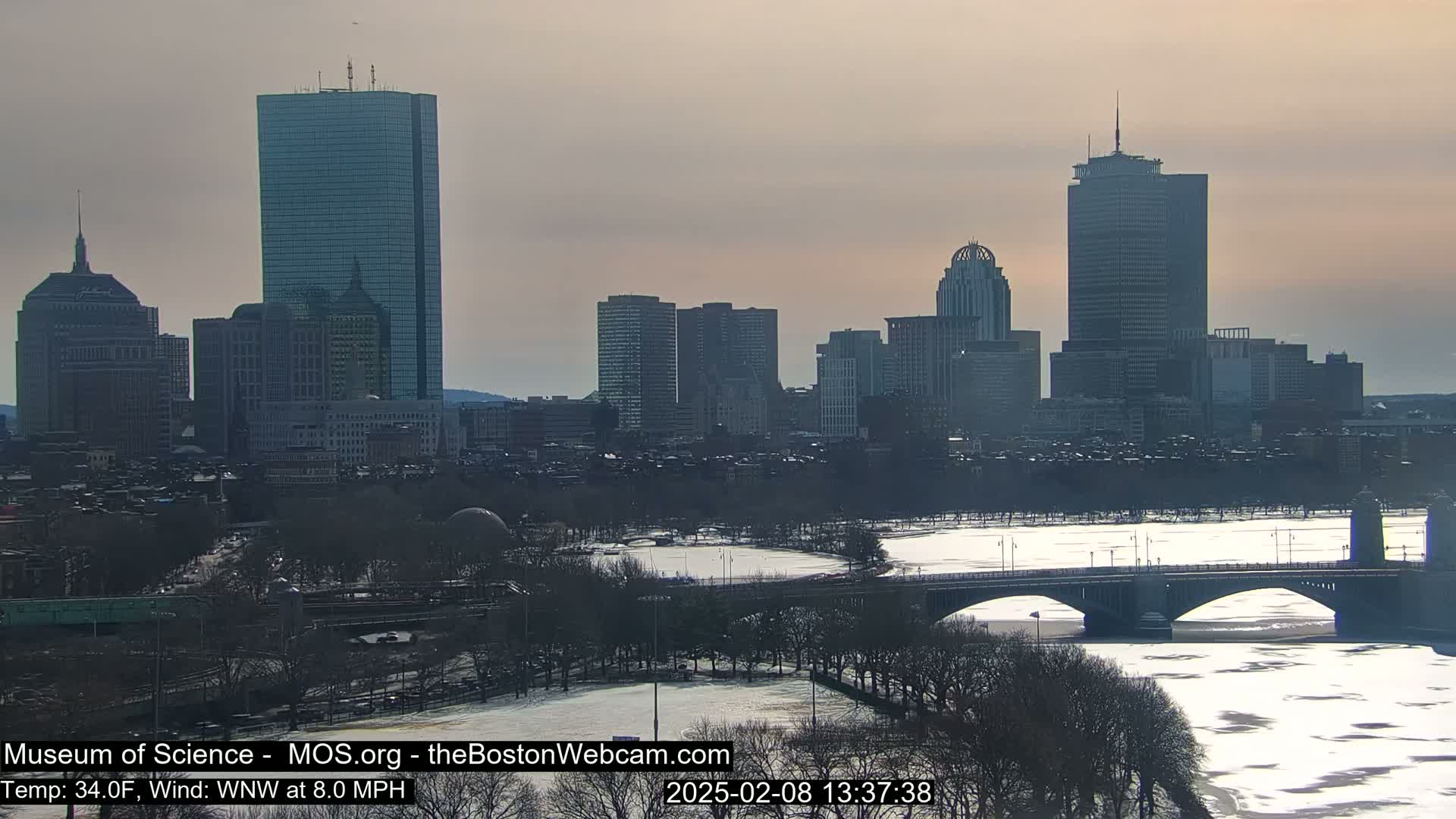 A city skyline with several tall buildings is visible across a partially frozen river, under a pale, overcast sky.