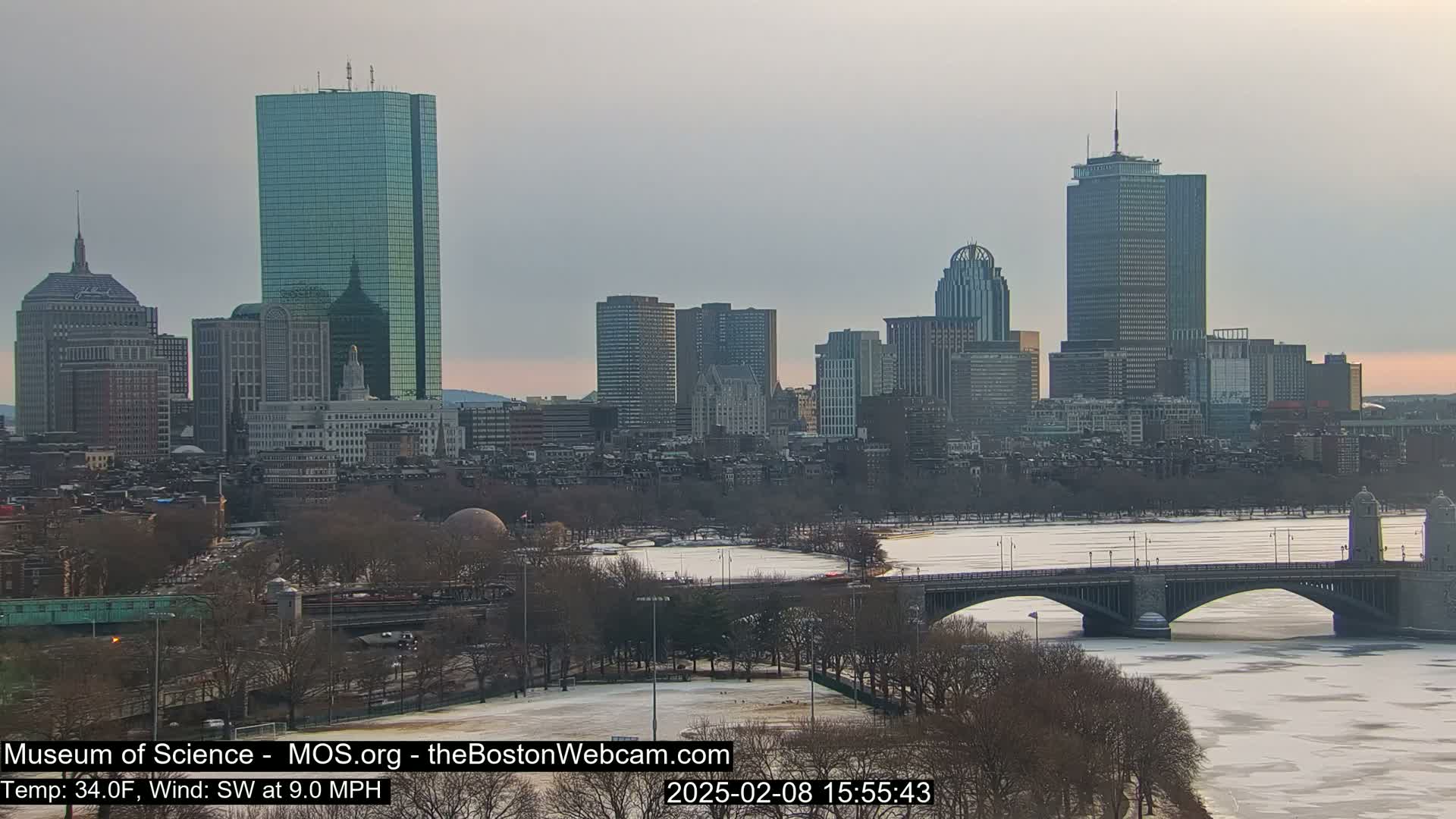A city skyline with several skyscrapers is seen across a partially frozen river and bridge under a partly cloudy sky.