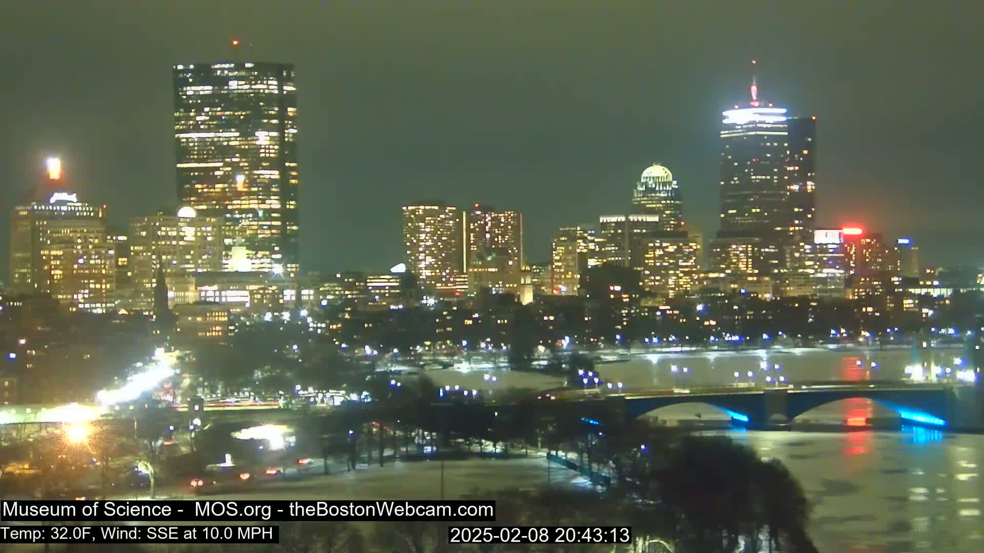 A nighttime view of a city skyline illuminated with lights, showing a bridge over a partially frozen river under a dark, clear sky with a southeasterly wind.