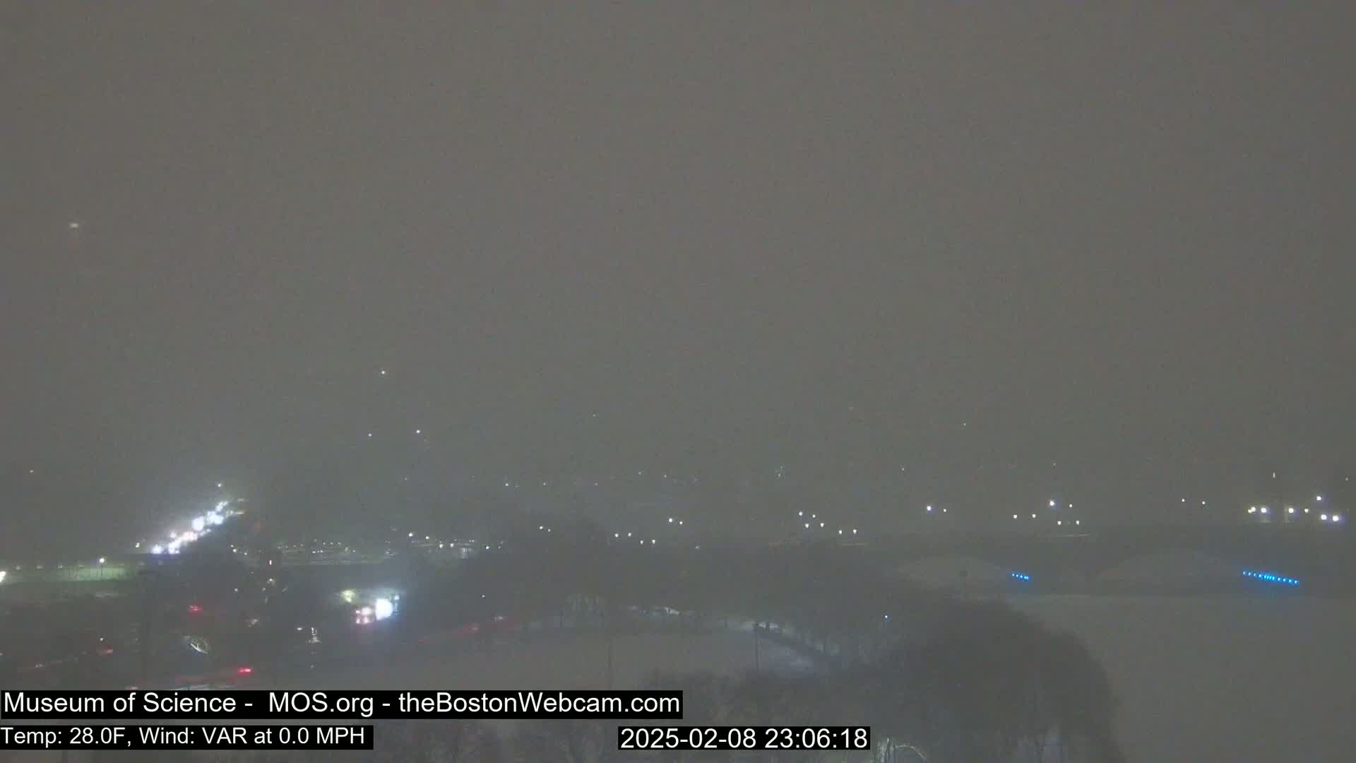 A hazy, nighttime view of a city landscape under snowy conditions, with dimly lit roads and buildings barely visible through the heavy snowfall.