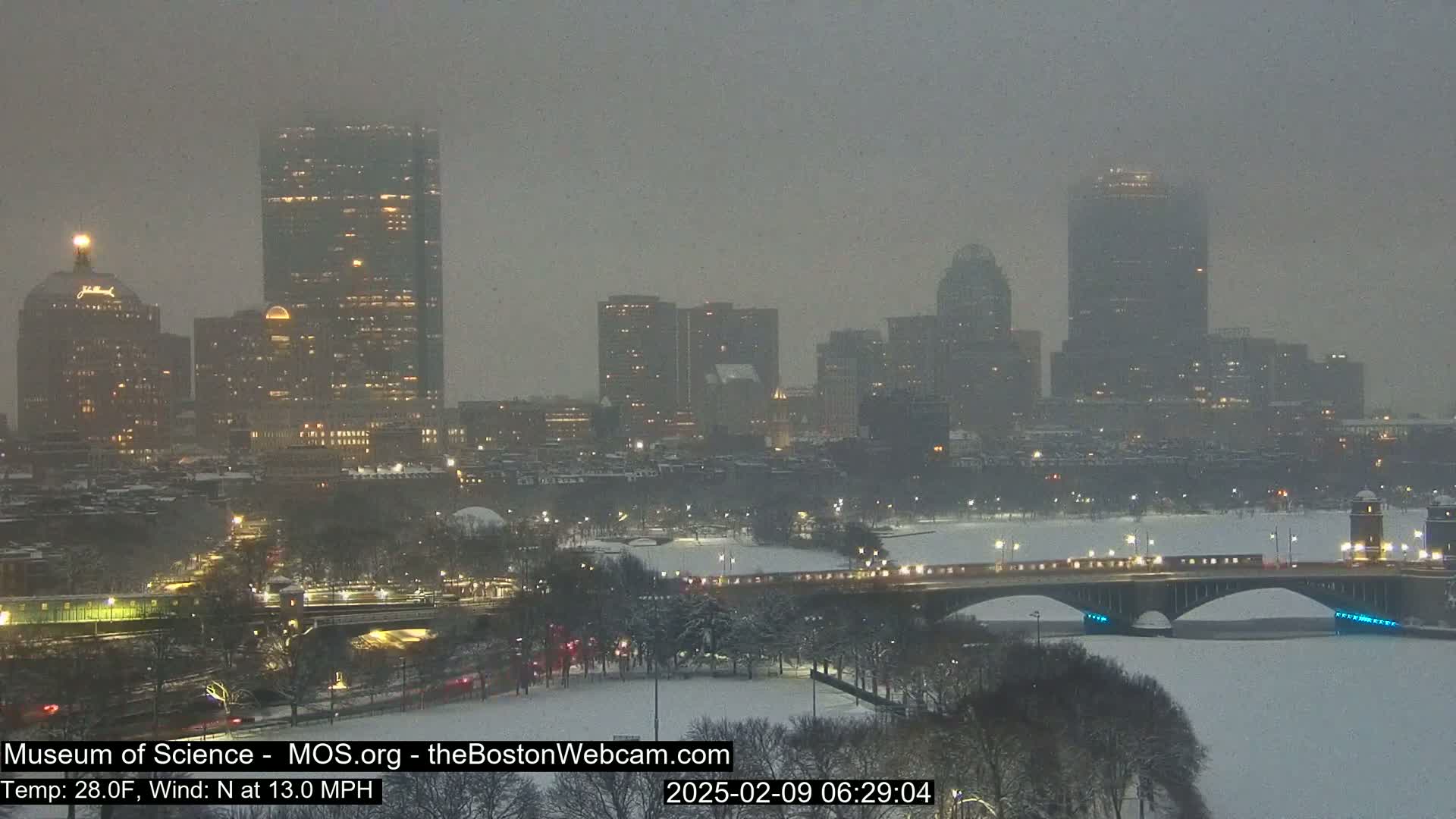 A snow-covered city skyline with illuminated buildings is seen across a river, with a bridge spanning the waterway under a light snowfall.