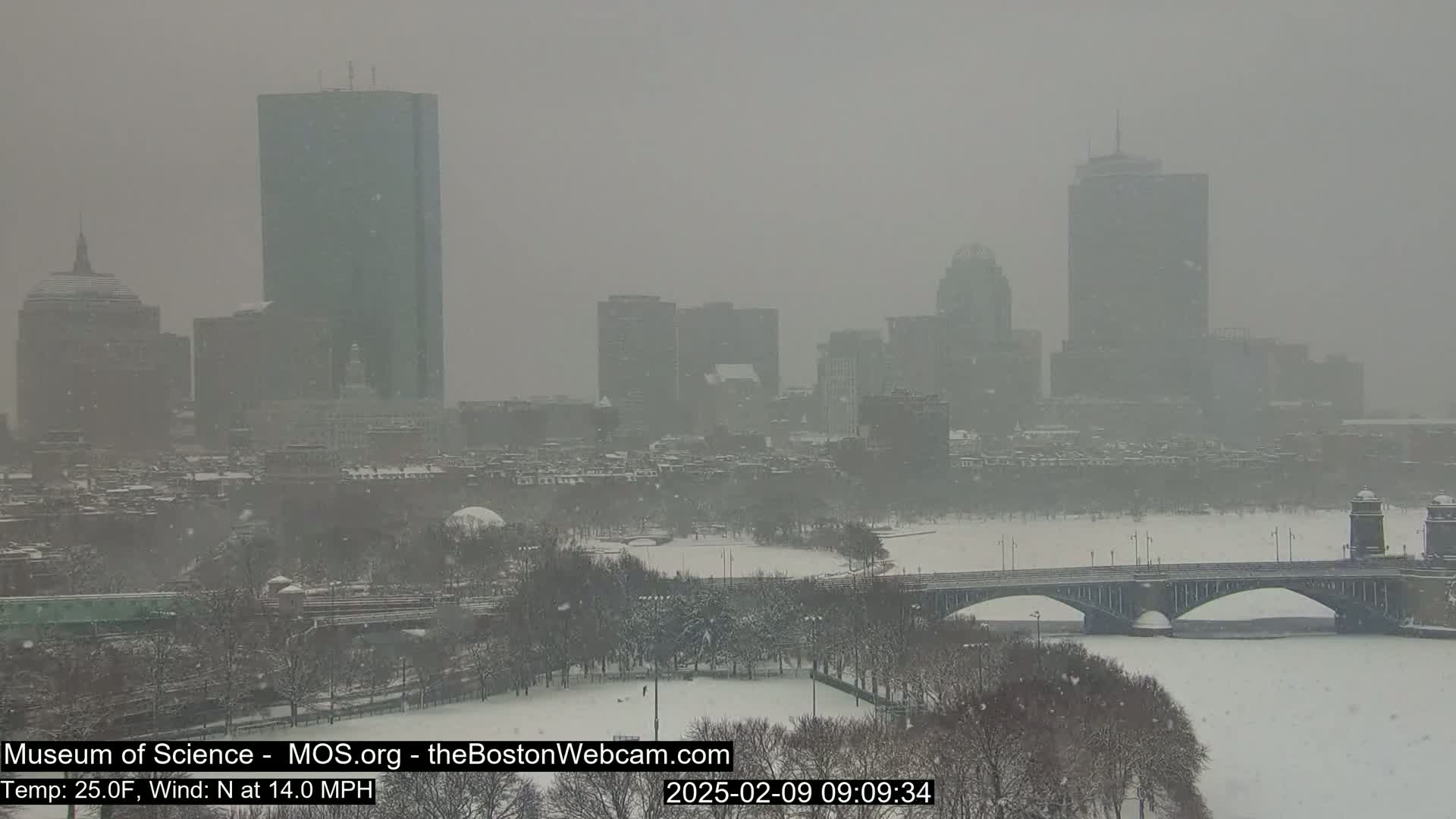 A snow-covered cityscape with a bridge over a frozen river is seen under a heavy snowfall.