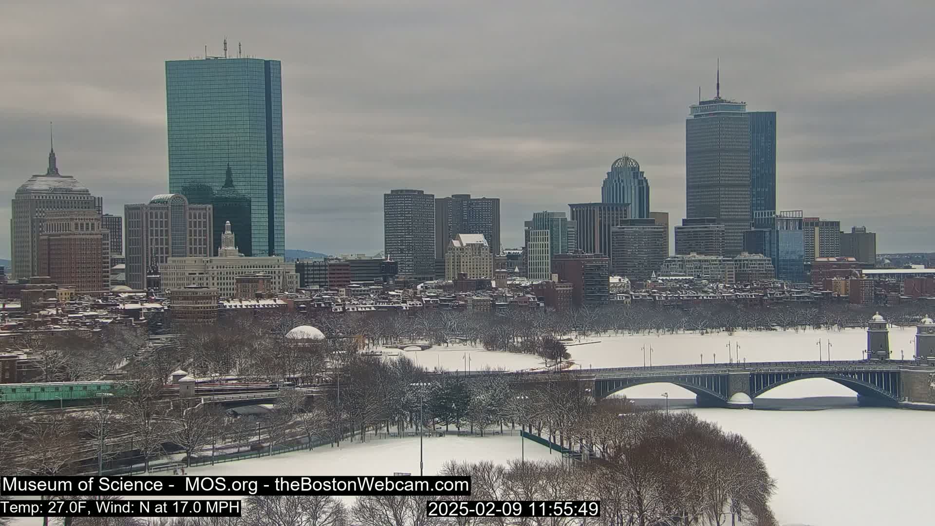 A snow-covered Boston cityscape, viewed from across a frozen river with a bridge, under an overcast sky.