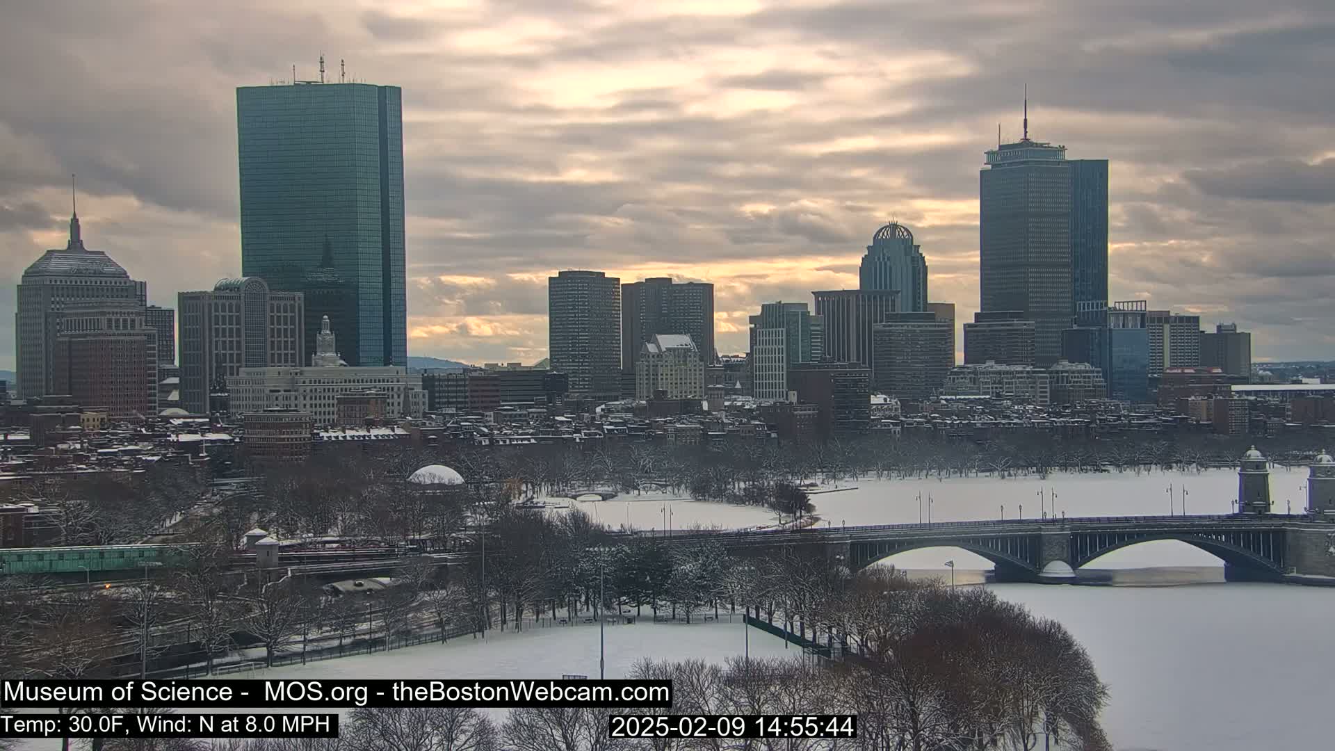 A snow-covered cityscape, with a river and bridge in the foreground, under a cloudy, dusky sky.