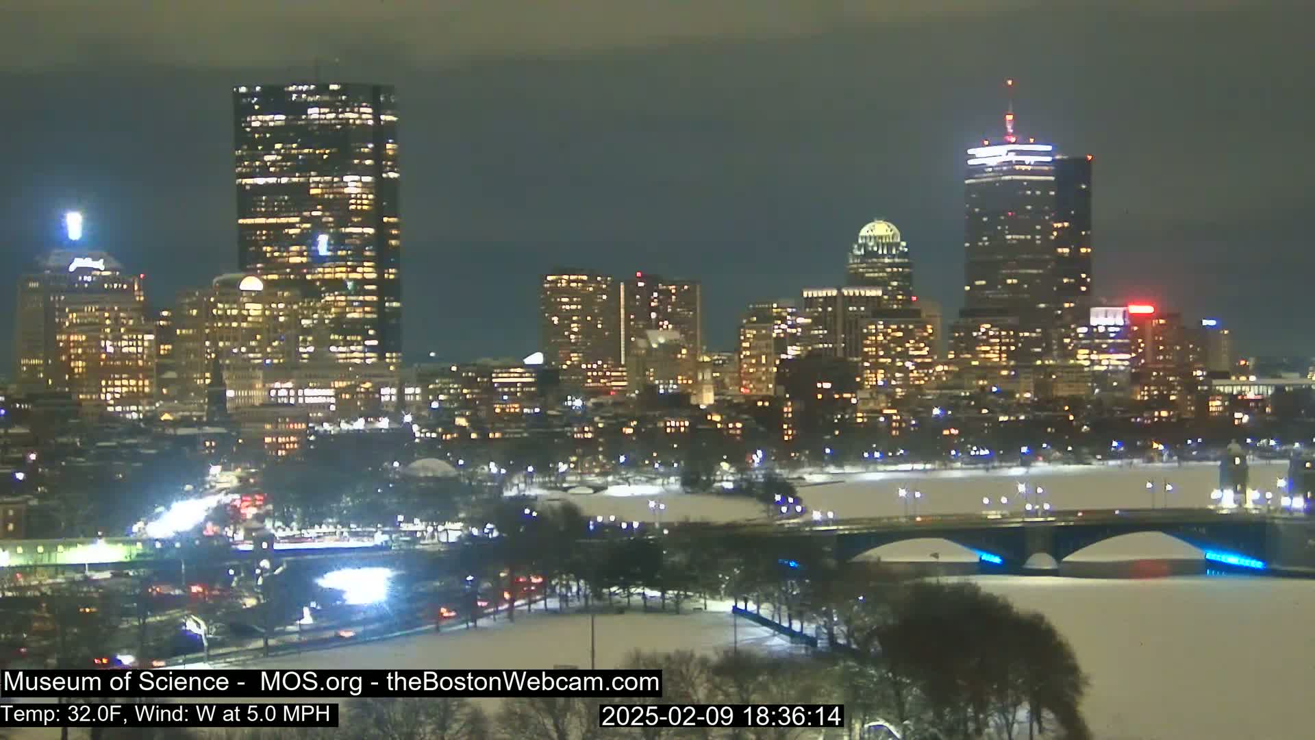 A nighttime view of a snow-covered city skyline with illuminated buildings and a bridge across a frozen river, under a dark, cloudy sky.