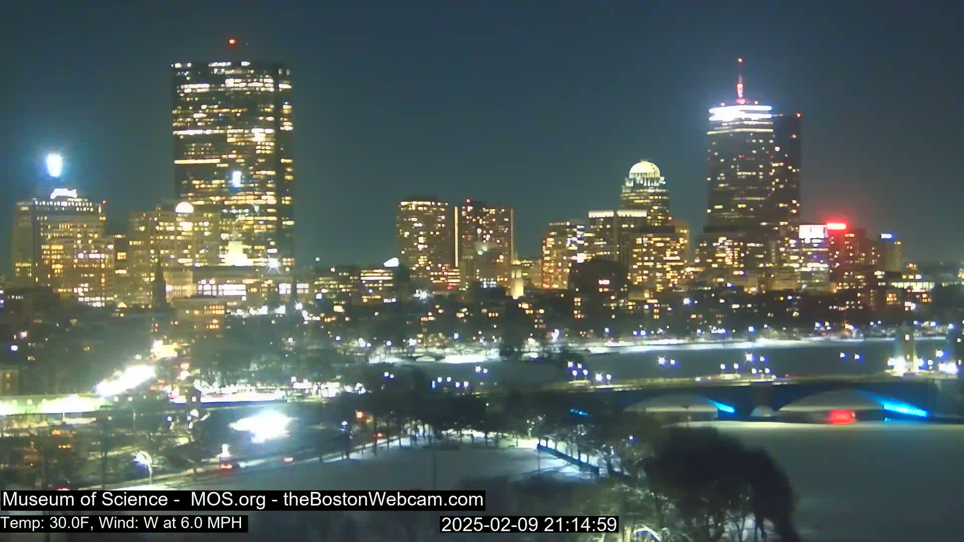 A nighttime view of a snow-covered city skyline with illuminated skyscrapers and a bridge over a partially frozen river under a clear, dark sky.