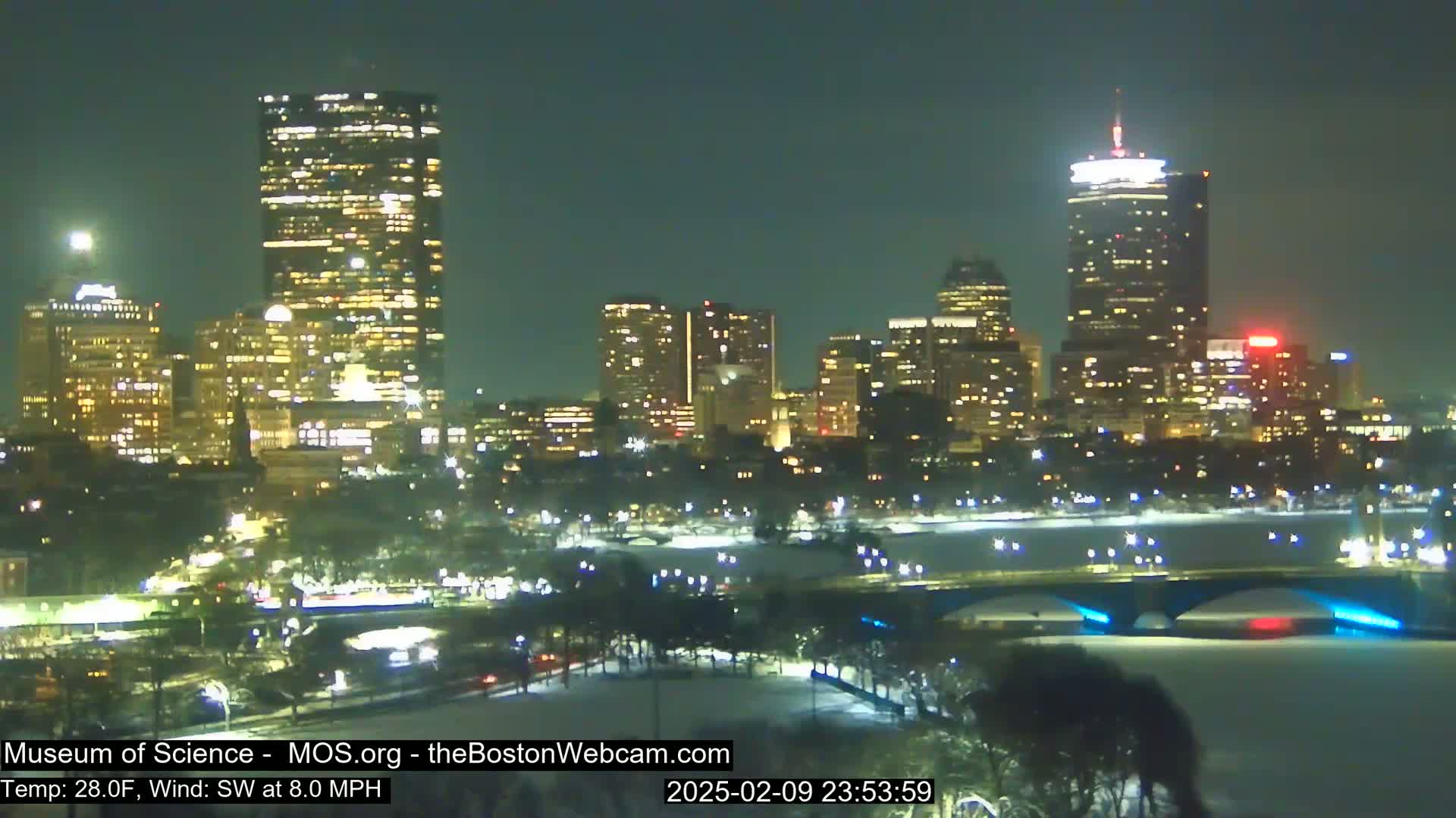 A nighttime view of a snow-covered city skyline with illuminated buildings and a bridge across a partially frozen river, under a clear sky.