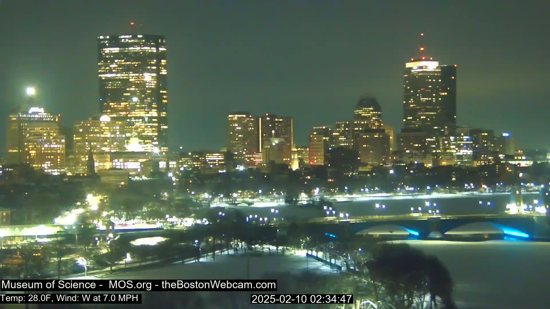 A nighttime view of a snow-covered city skyline illuminated by building lights, with a bridge crossing a partially frozen body of water under a clear sky.