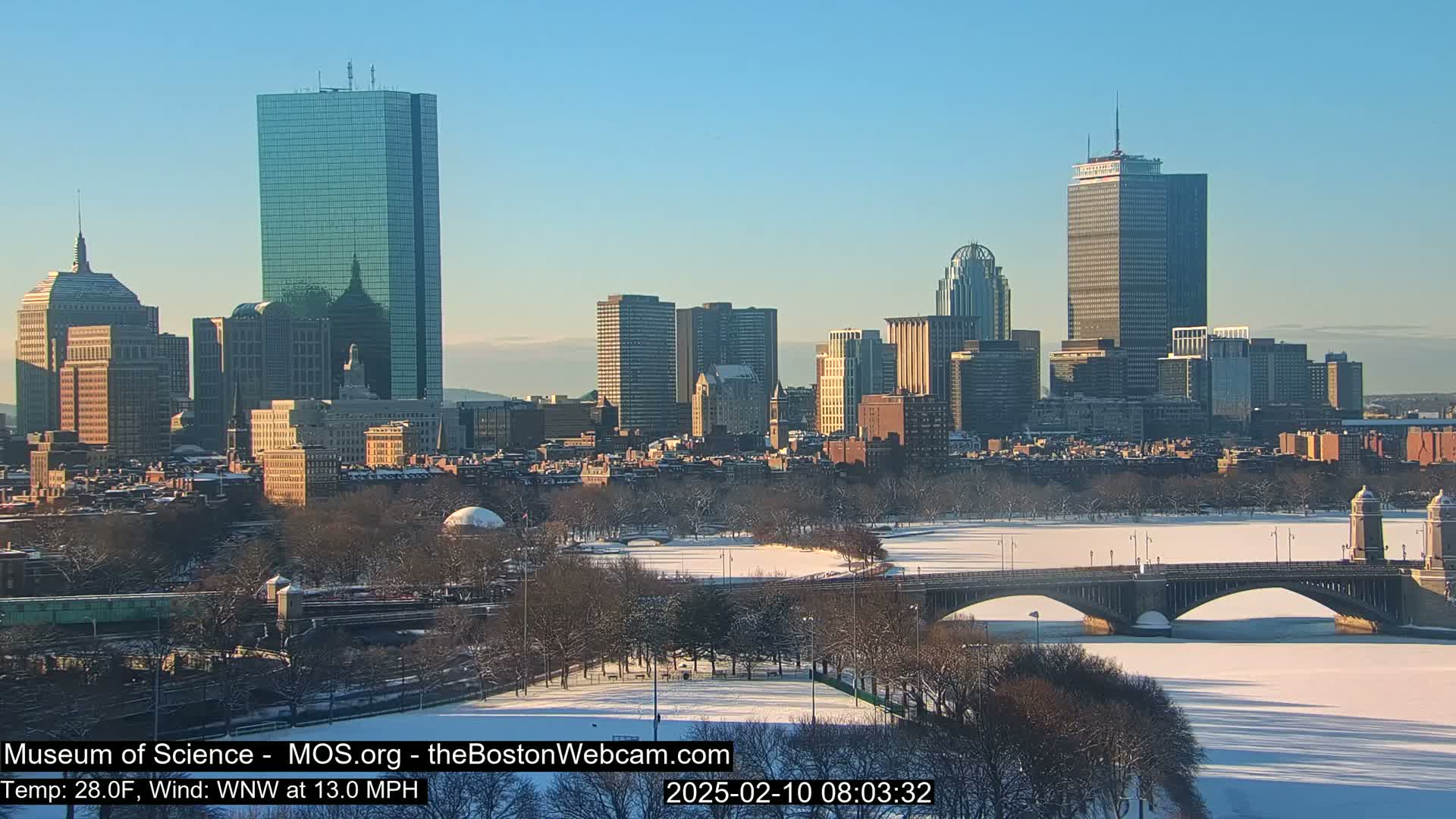 A snow-covered city skyline with a frozen river and bridge underneath a clear, sunny sky.