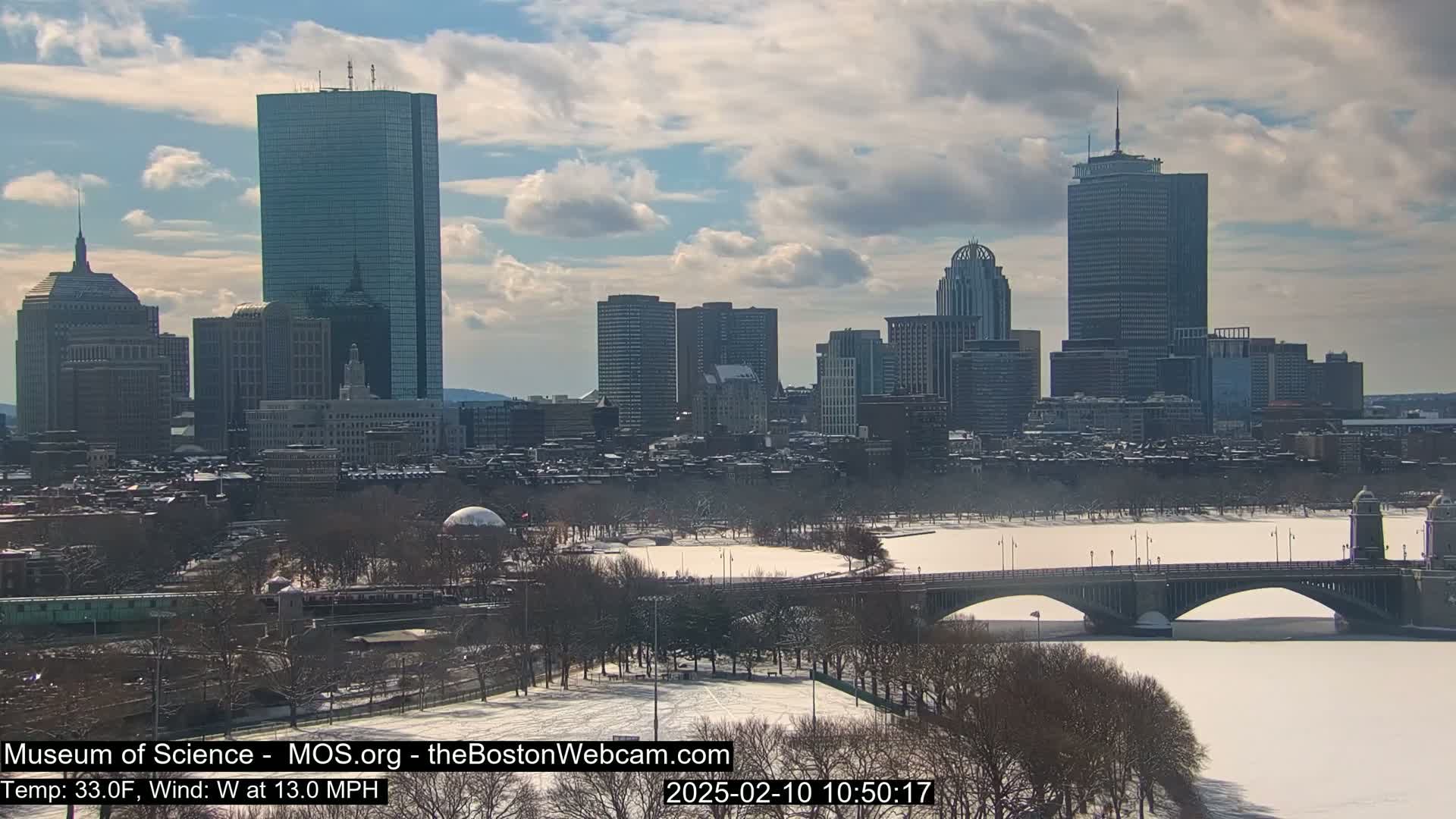 A snow-covered city skyline with a partially frozen river and a bridge in the foreground under a partly cloudy sky.