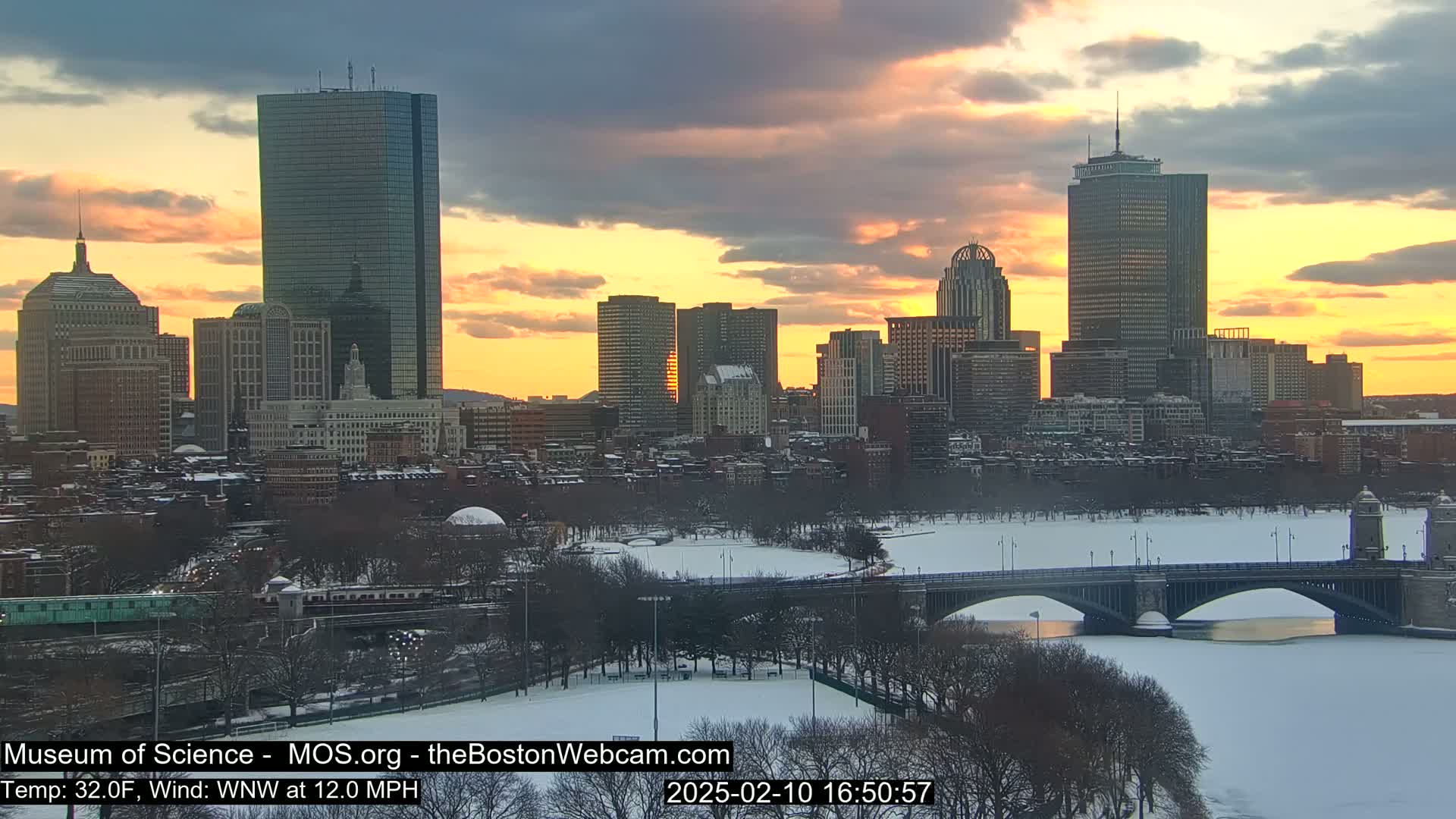 A snow-covered cityscape at sunset, with a bridge spanning a partially frozen river in the foreground.