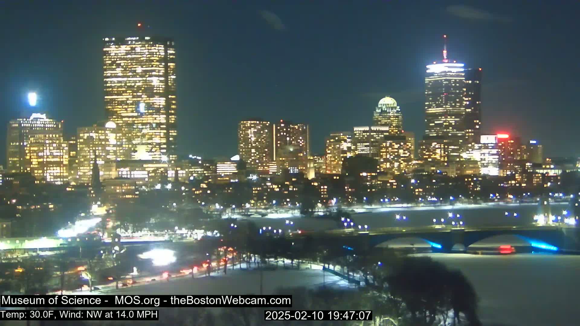 A nighttime view of a city skyline illuminated with lights, under a clear sky, with a river and bridge in the foreground and snow on the ground.