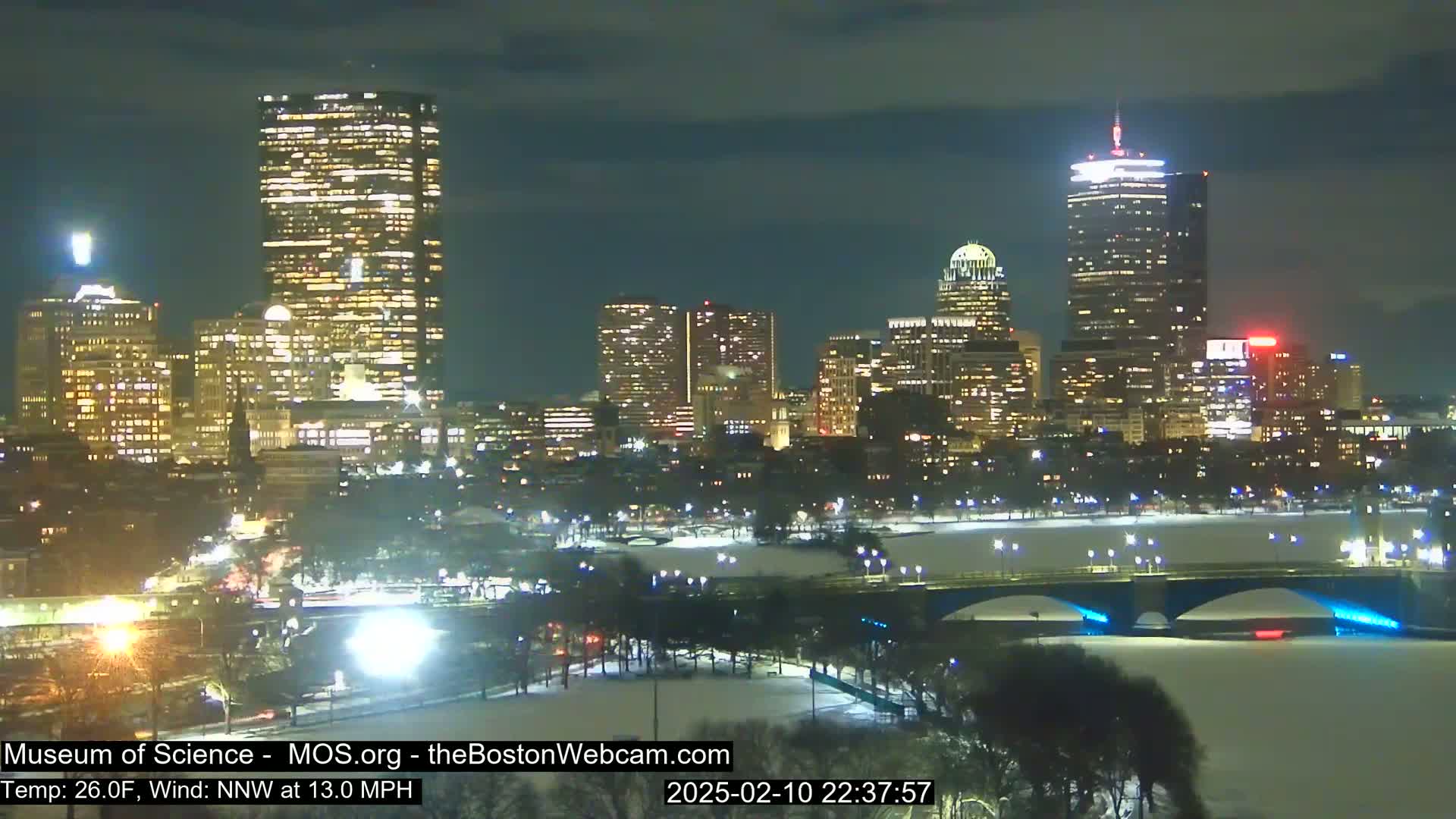 A nighttime cityscape with snow-covered ground and a bridge illuminated, under a dark, mostly clear sky.