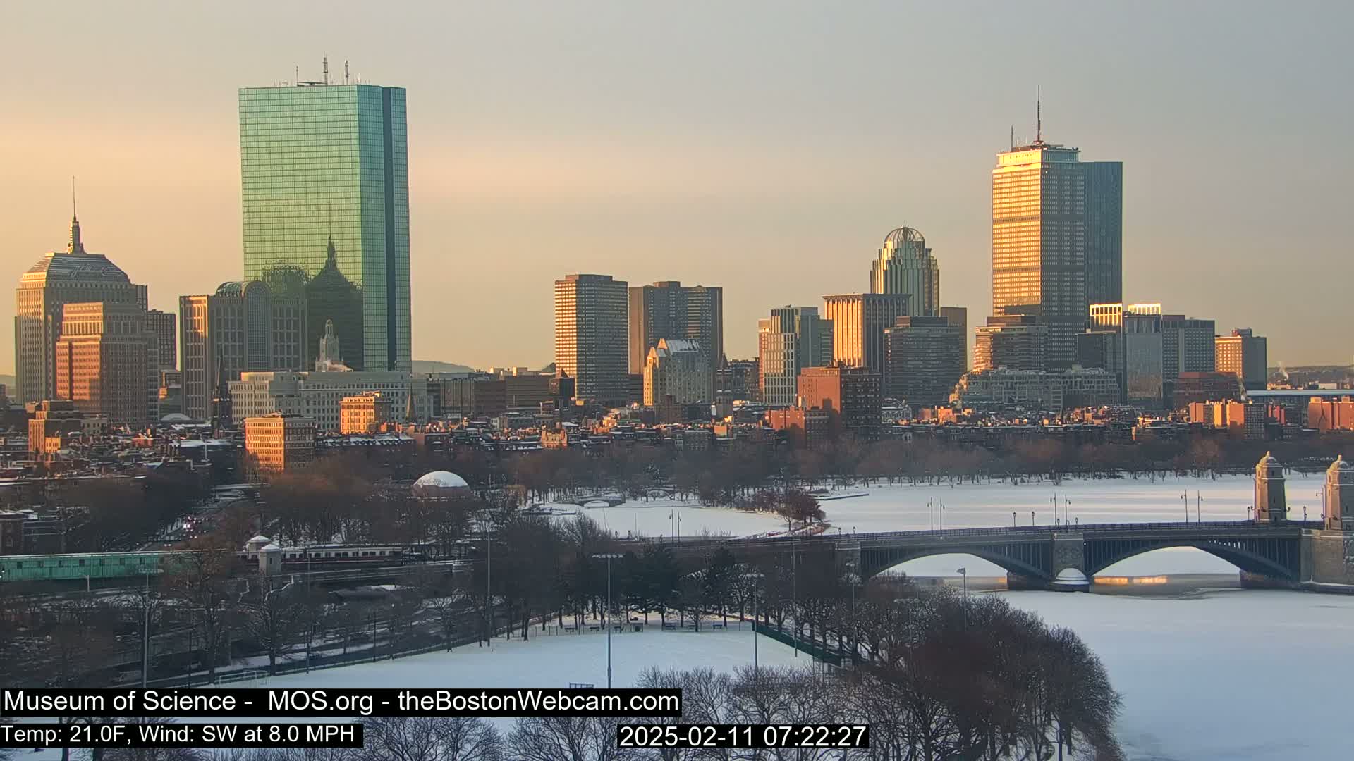 A snow-covered city skyline with a bridge over a frozen river is bathed in the warm light of sunrise.