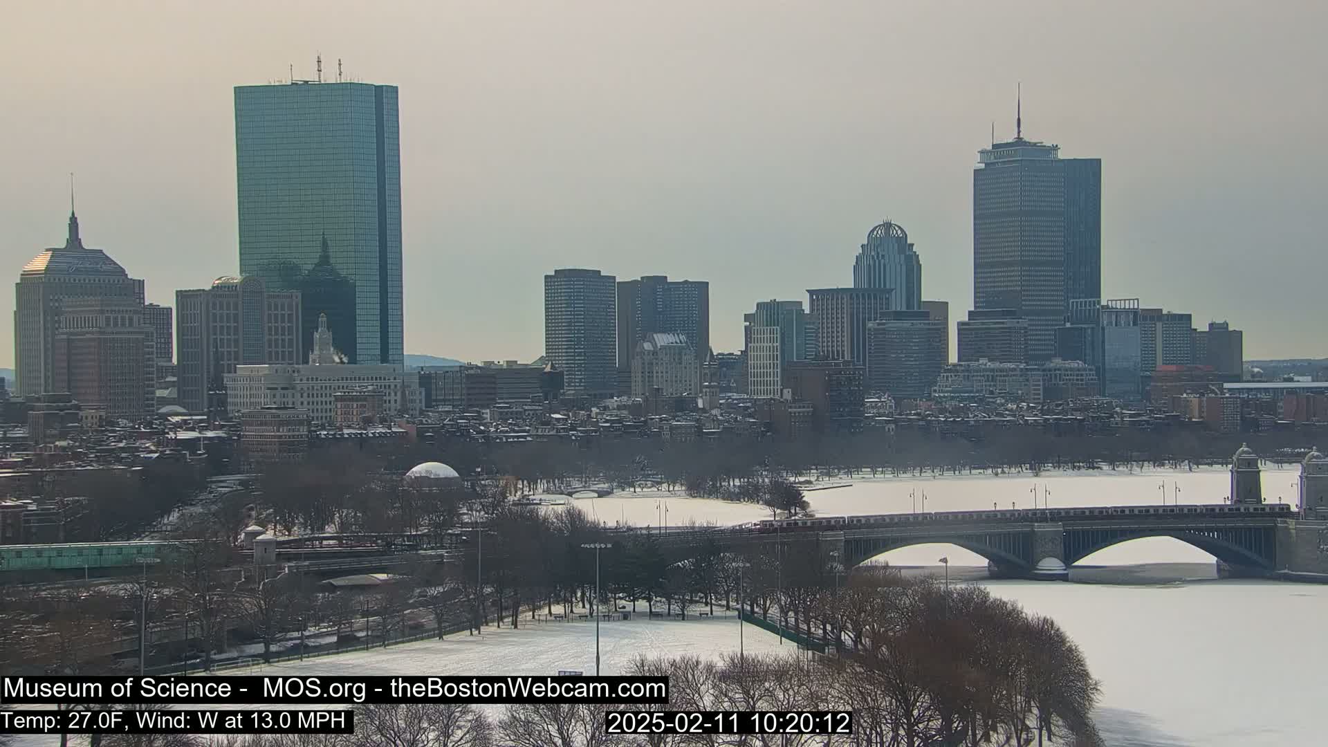 A snow-covered city skyline, including several tall skyscrapers, is viewed from across a frozen river with a bridge in the foreground.