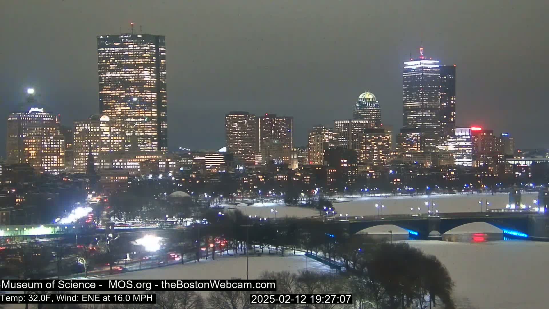 A nighttime view of a snow-covered city skyline with brightly lit skyscrapers and a bridge over a partially frozen river, under a dark, overcast sky with a 32°F temperature and 16 mph east-northeast wind.