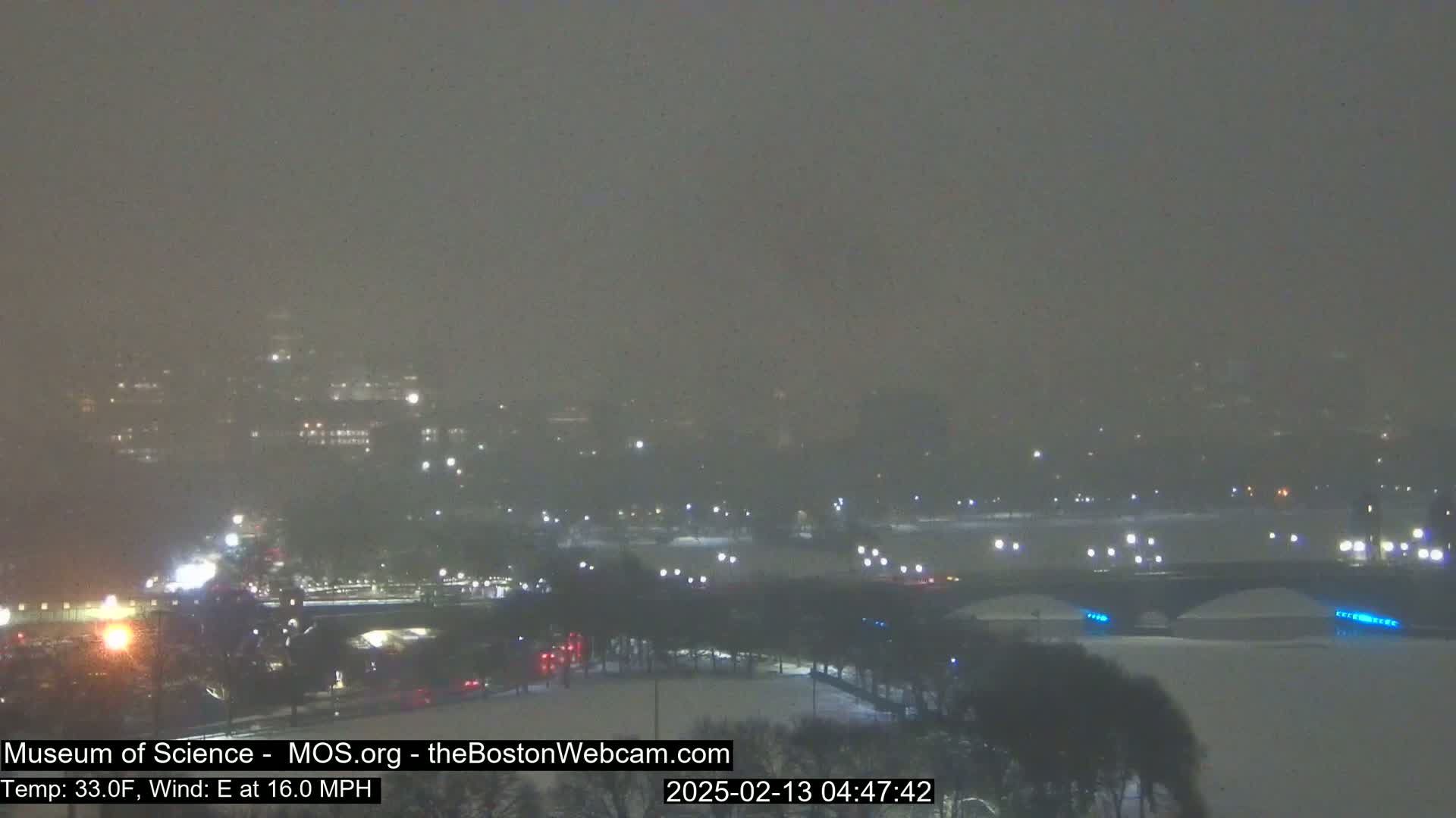A snowy, nighttime scene shows a bridge over a river, with city lights and buildings dimly visible in the distance through heavy snowfall and blowing snow.