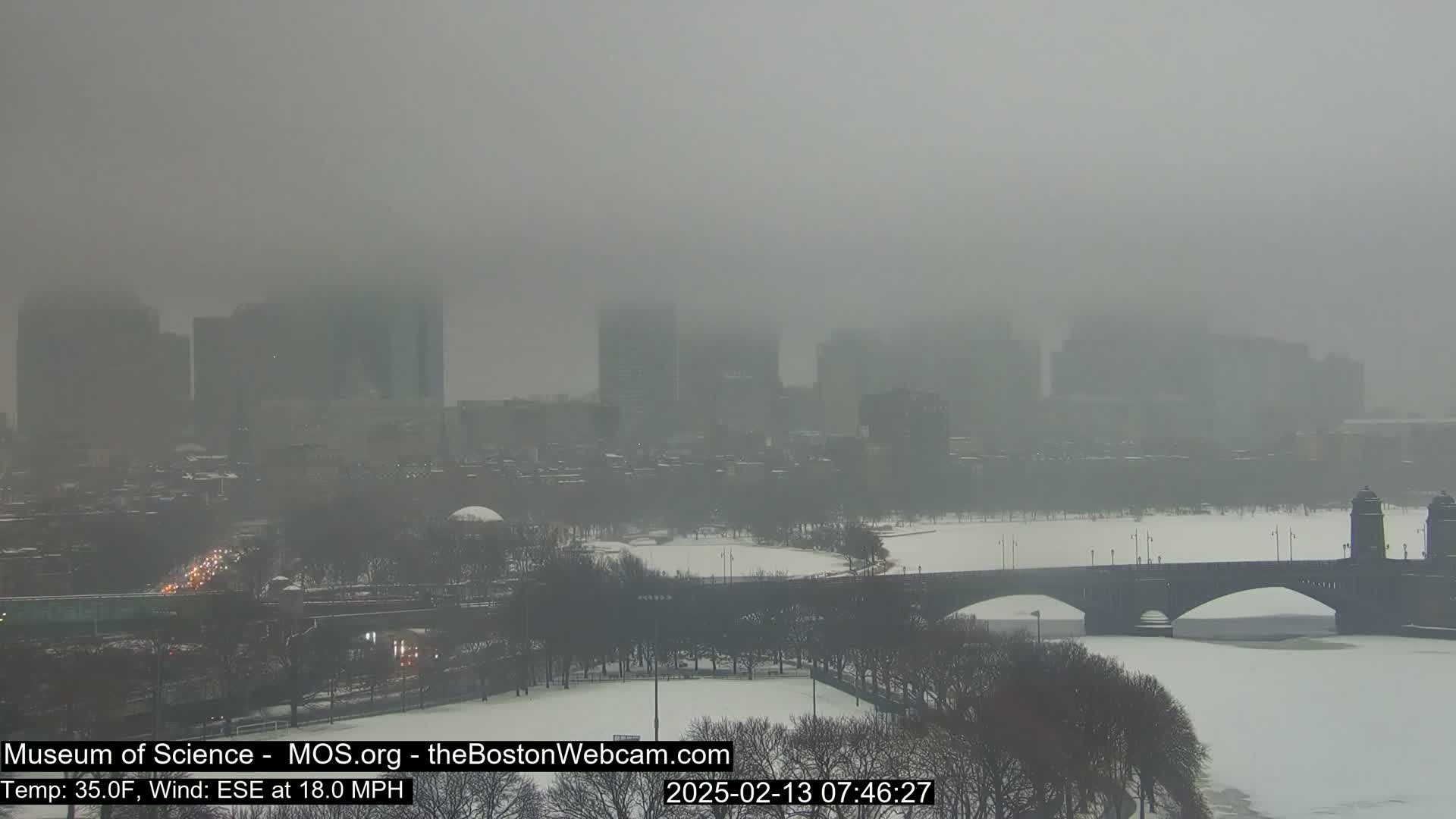 A hazy, snowy cityscape is visible across a frozen river and bridge, with a strong wind.