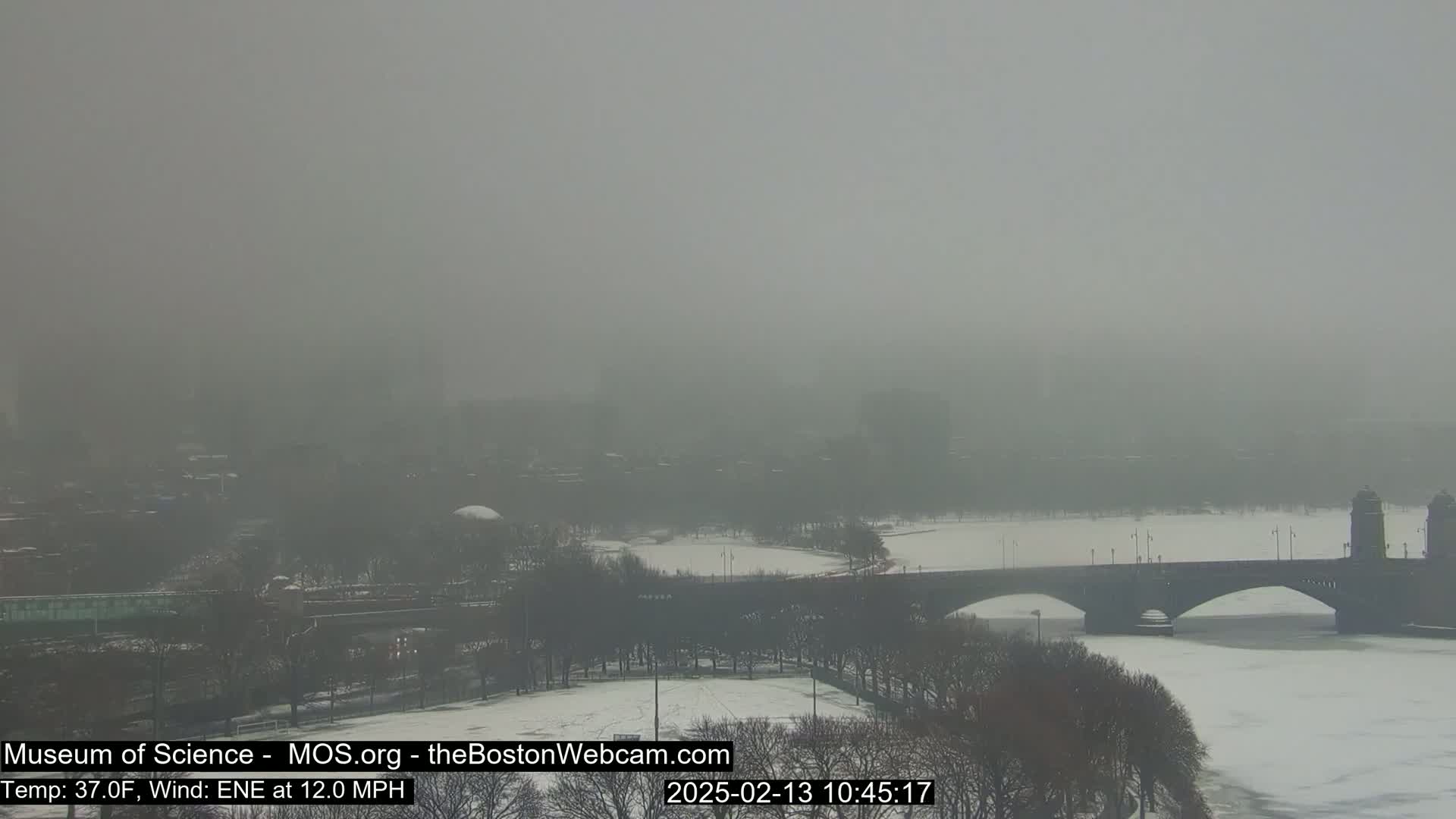 A snow-covered bridge spans a foggy river, with a hazy cityscape visible in the background under a gray sky.