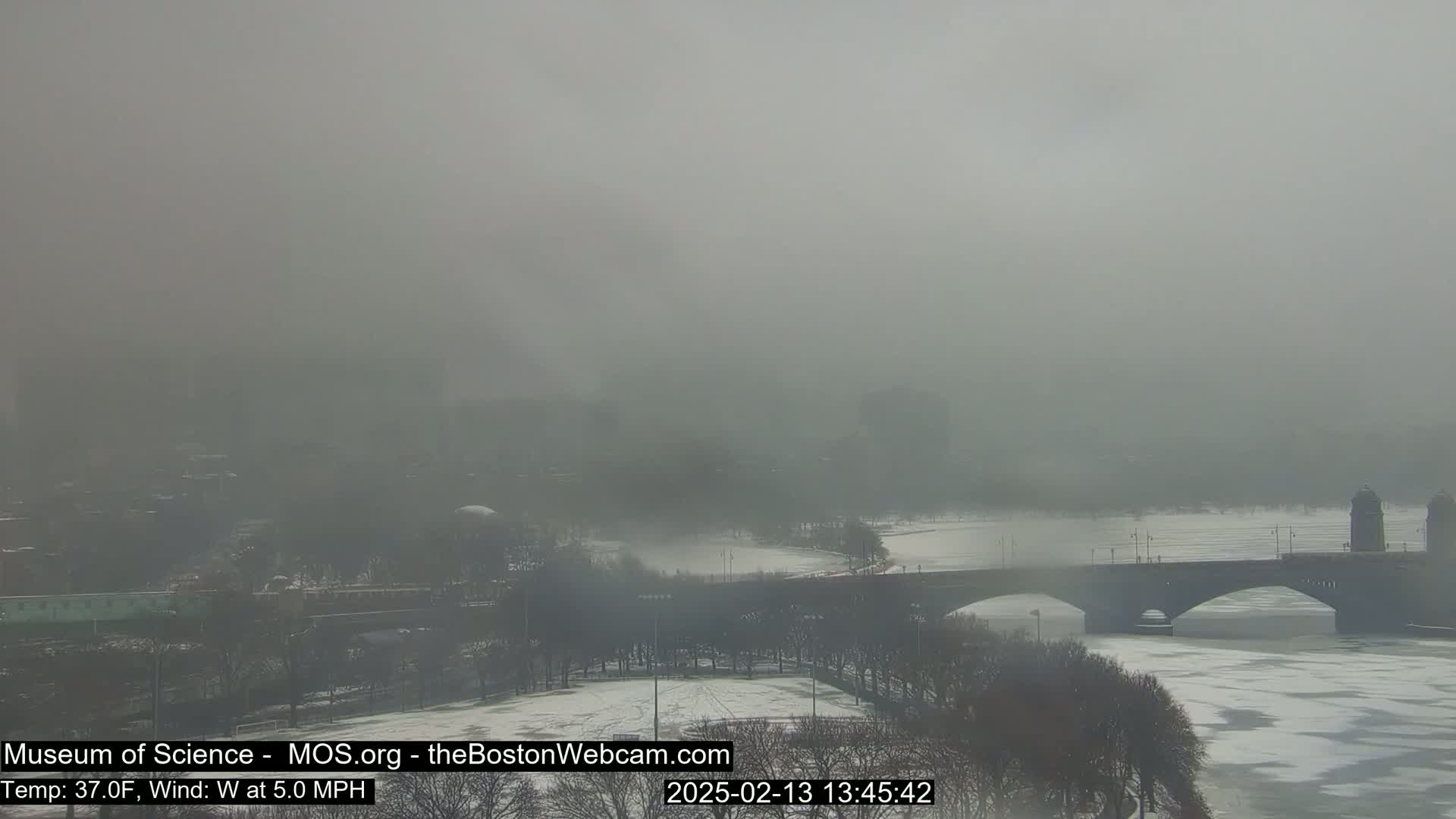A foggy, wintry scene shows a bridge spanning a partially frozen river, with trees and buildings barely visible in the distance.