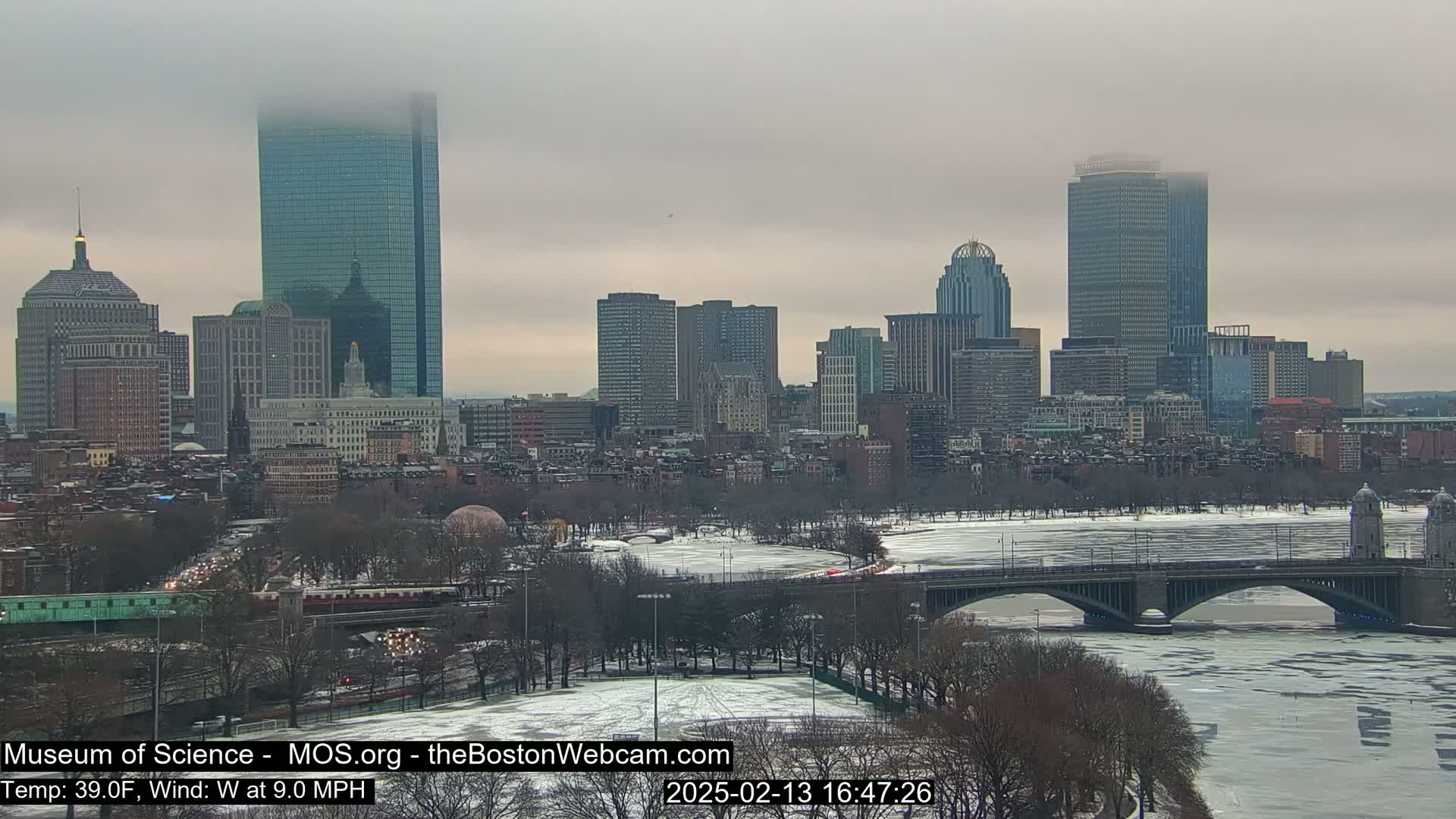 A city skyline with several skyscrapers is visible under an overcast sky, with a river partially covered in ice in the foreground.