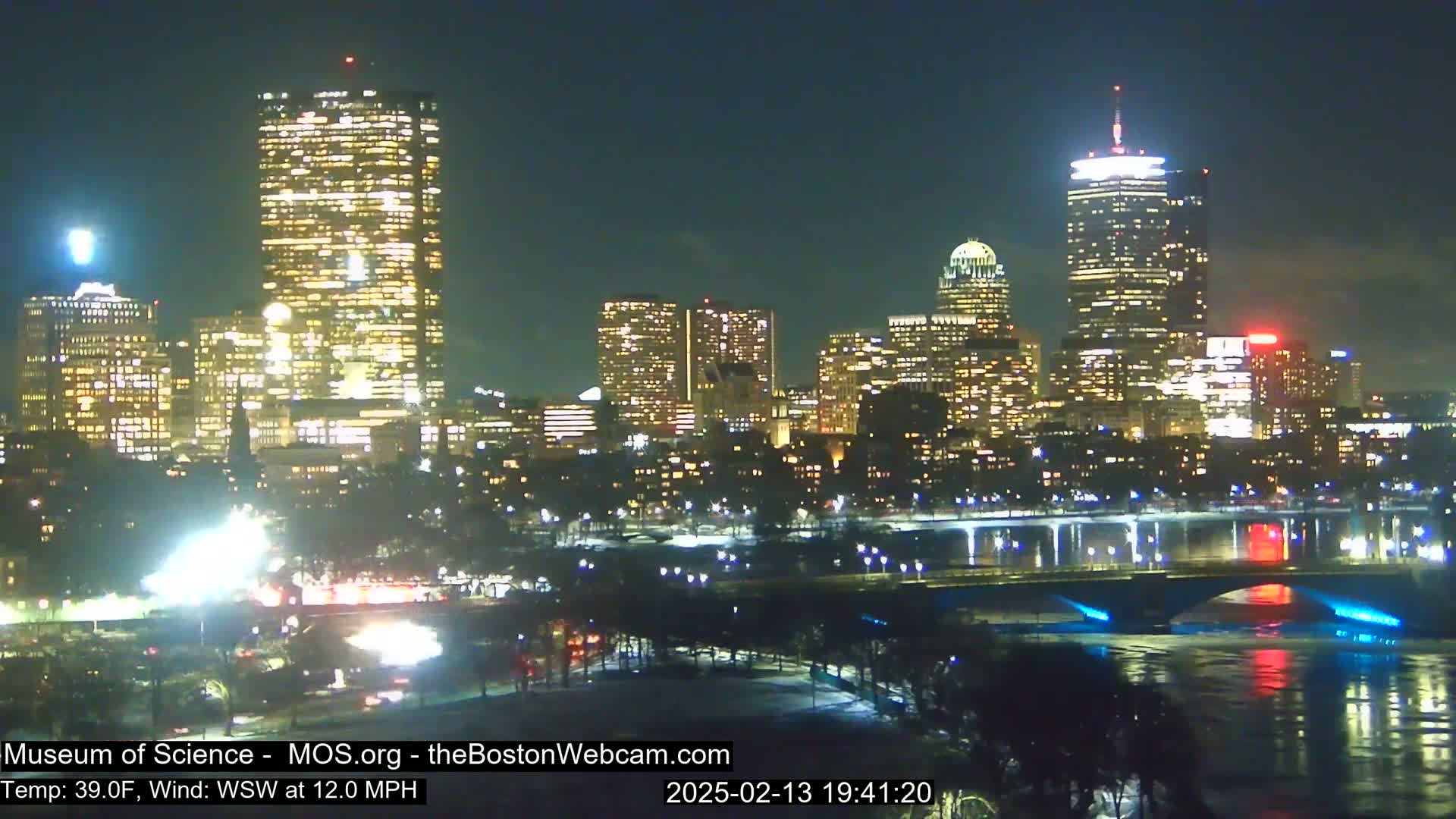 A nighttime view of a city skyline with numerous illuminated buildings, a bridge, and a body of water, under a partly cloudy sky with a wind of 12 mph from the west-southwest.