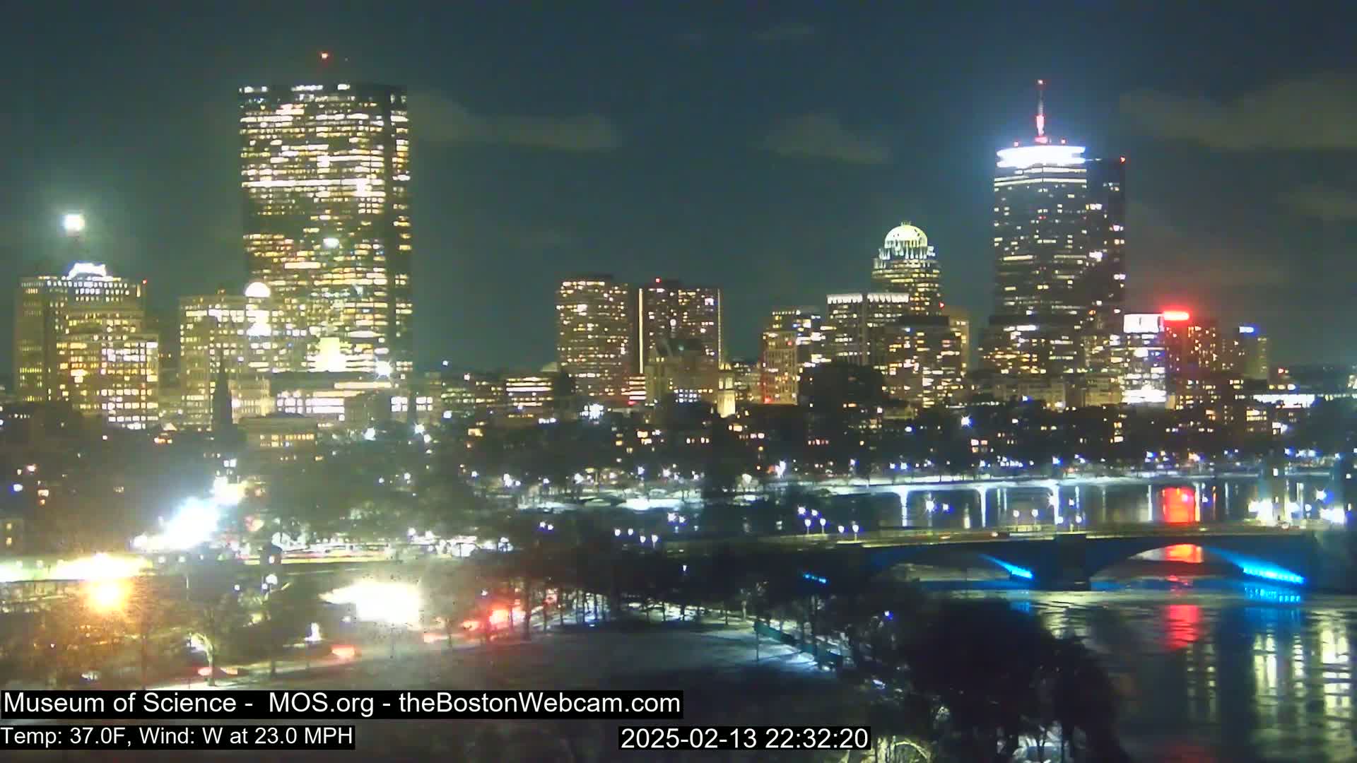 A nighttime view of a city skyline illuminated with bright lights, featuring several tall buildings and a bridge across a body of water under a partly cloudy sky, with a west wind at 23 mph and a temperature of 37°F.