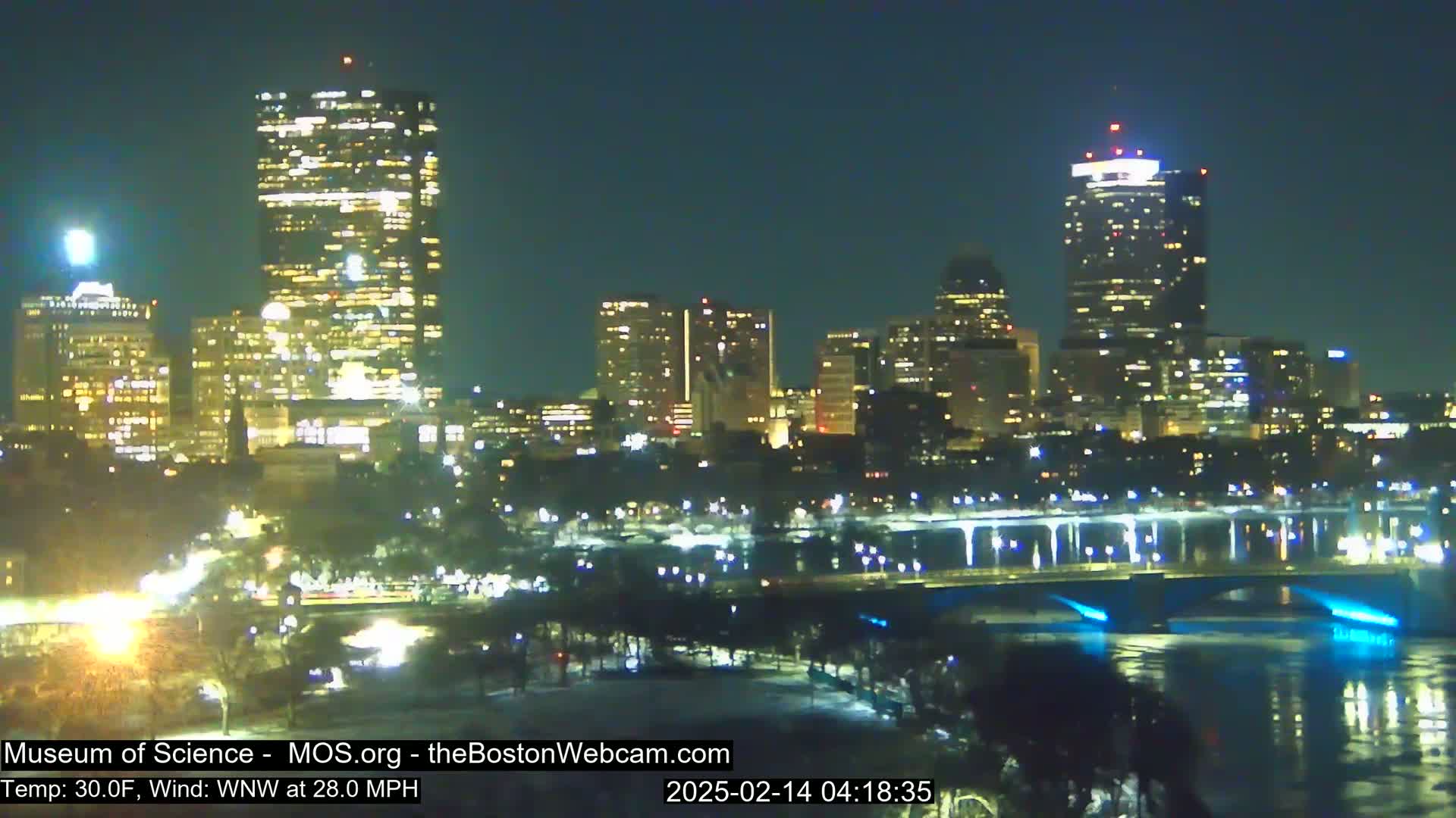 A nighttime cityscape with numerous illuminated buildings, a bridge, and a river, under a clear sky with a 30°F temperature and a 28 mph wind from the west-northwest.