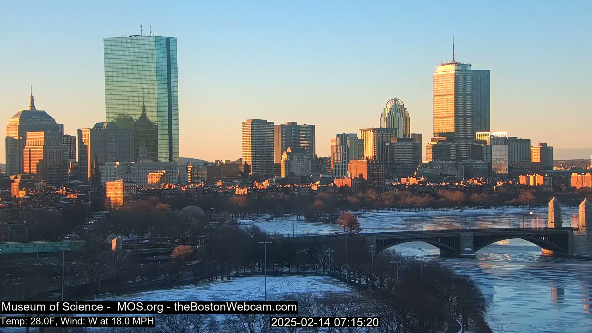 A city skyline at sunrise with a partially frozen river and bridge in the foreground, under a clear, cold sky.