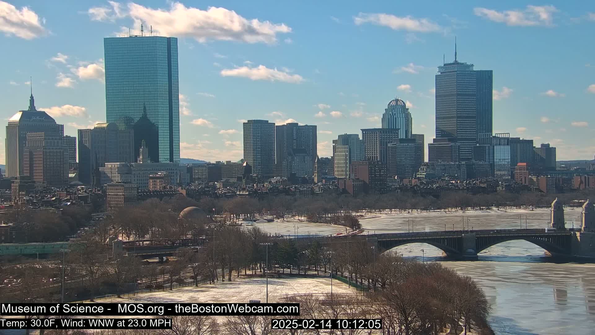 Under a partly cloudy, sunny sky, a city skyline is visible across a partially frozen river, with a bridge in the foreground and snow-covered parkland.