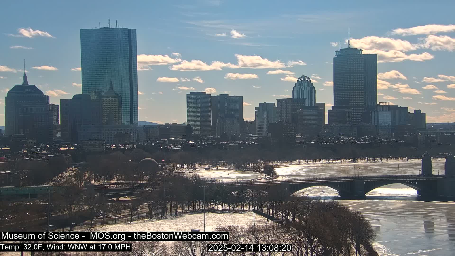 A partly cloudy day reveals a cityscape with numerous skyscrapers, a river partially frozen over, and a bridge spanning the water.