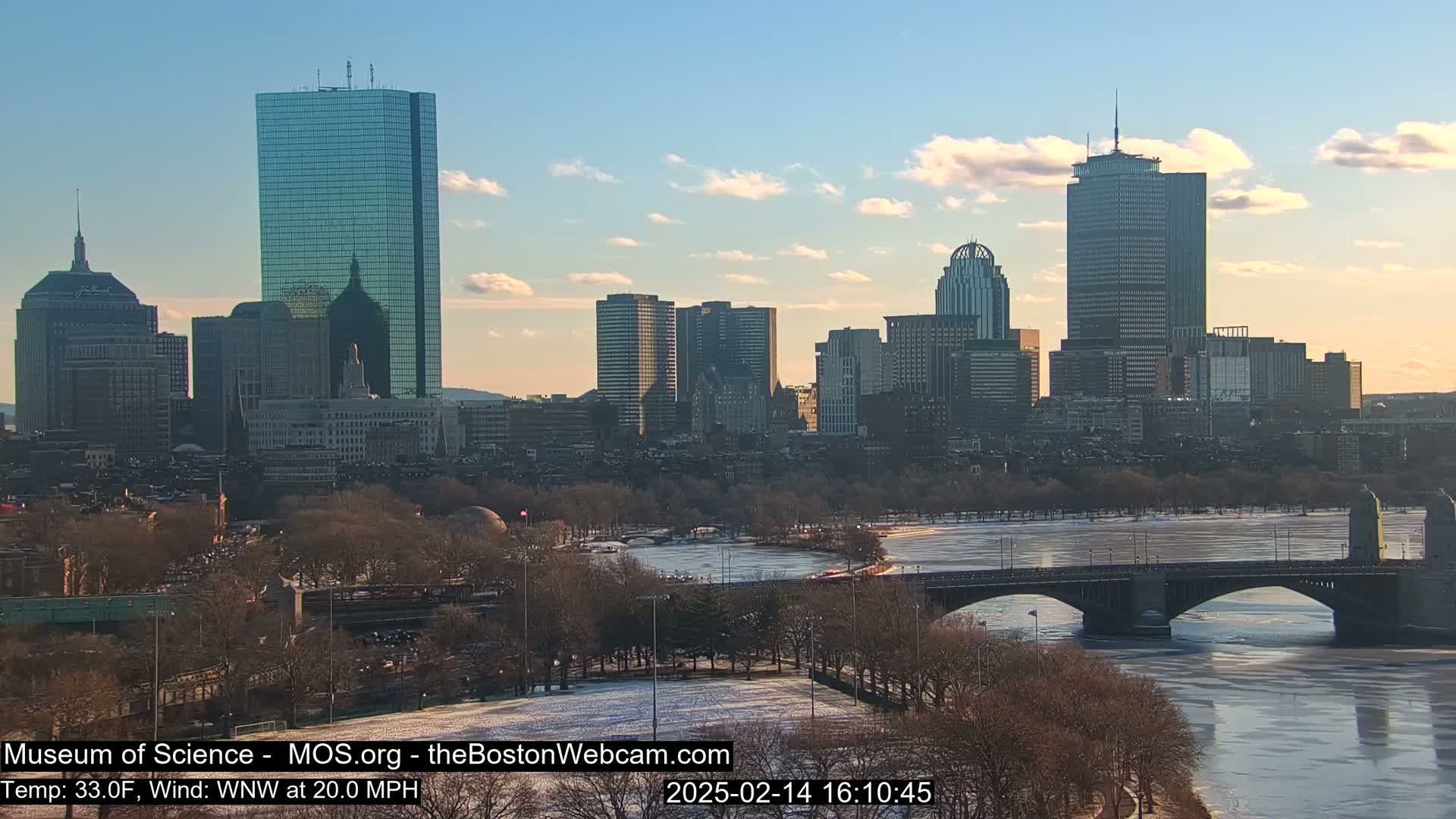 A city skyline with several skyscrapers is visible across a partially frozen river under a partly cloudy sky.