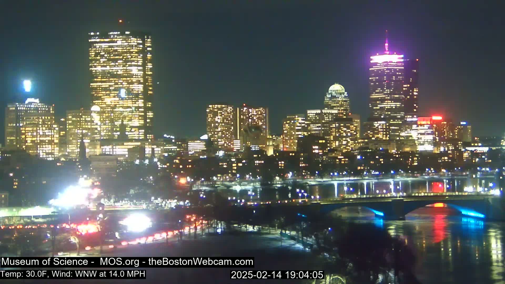 A nighttime view of a city skyline illuminated with numerous lights, featuring a river with a bridge and traffic visible on a roadway in the foreground.