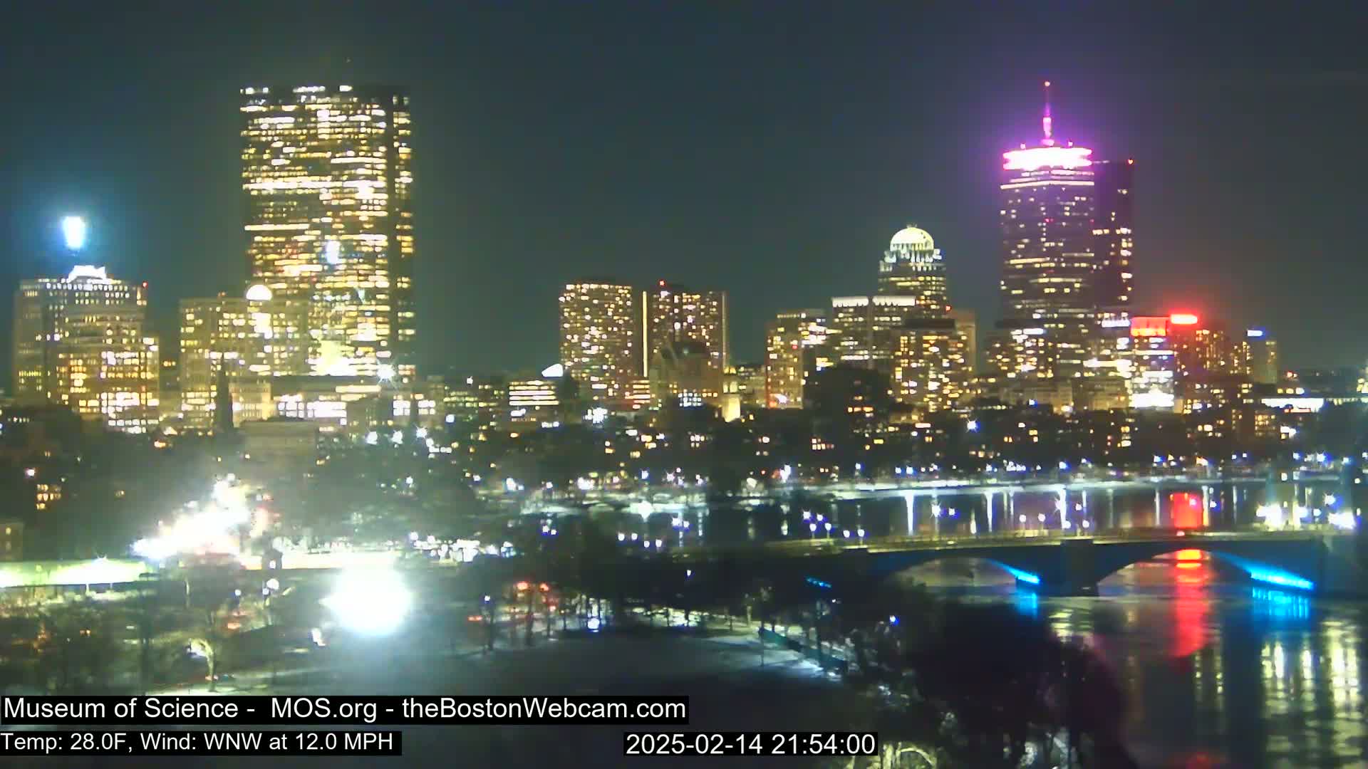 A brightly lit city skyline at night is seen across a dark river, with a bridge illuminated in blue and red lights, under a clear night sky.