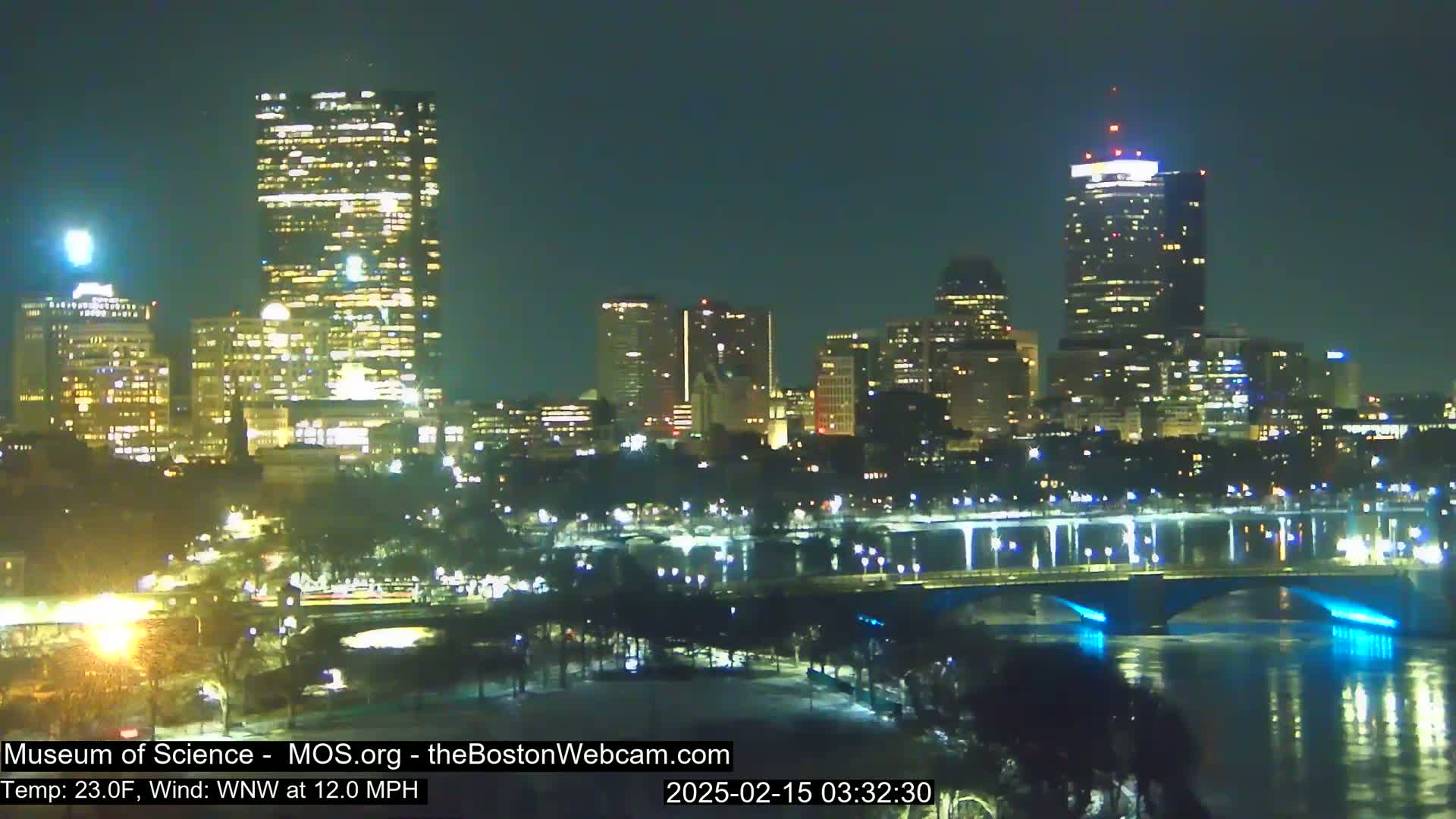A nighttime view of a city skyline with illuminated buildings across a river, under a clear sky with a 23°F temperature and a 12 mph wind from the west-northwest.