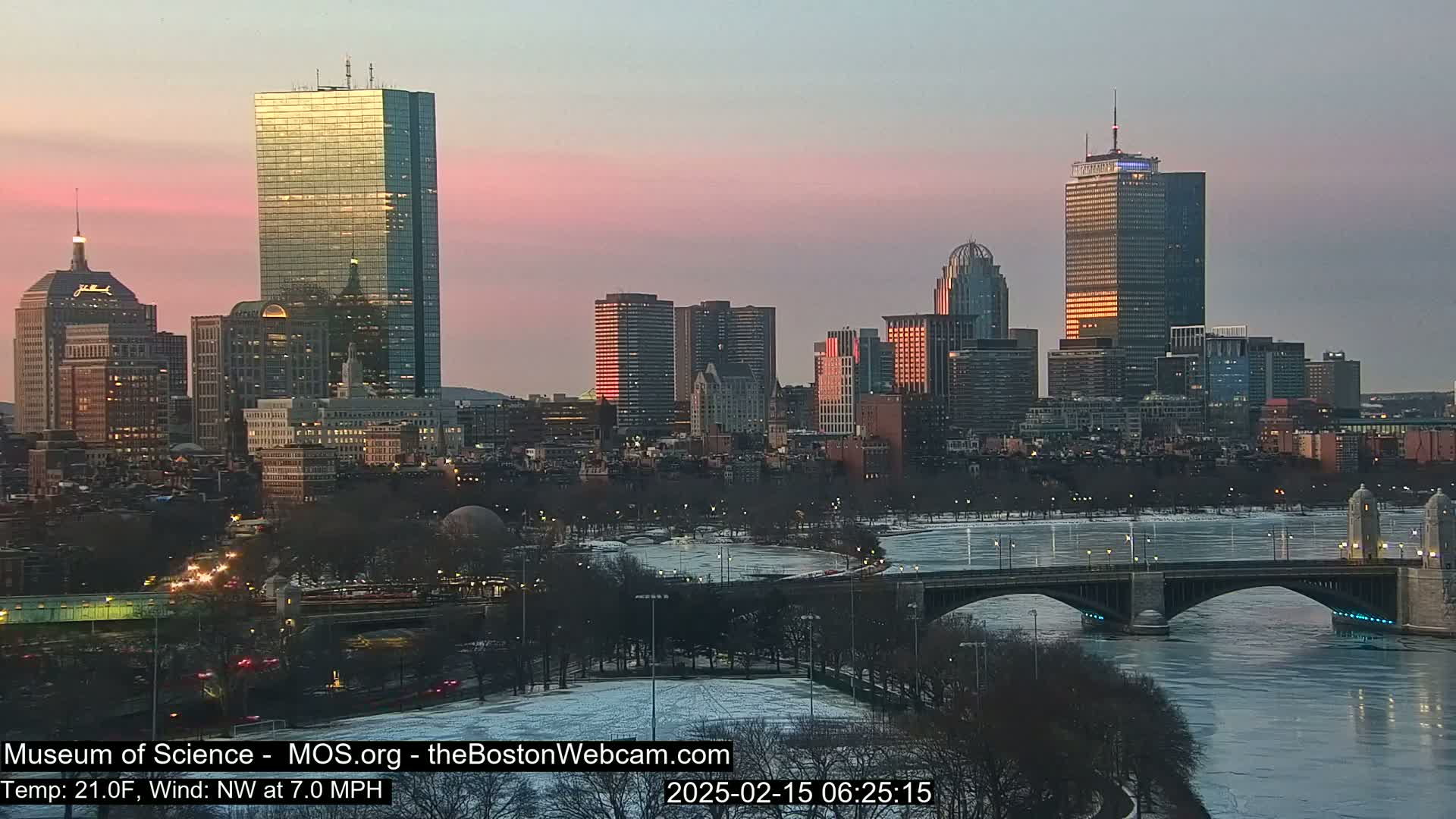 A city skyline at sunrise with a partially frozen river and bridge in the foreground, under a pinkish sky.