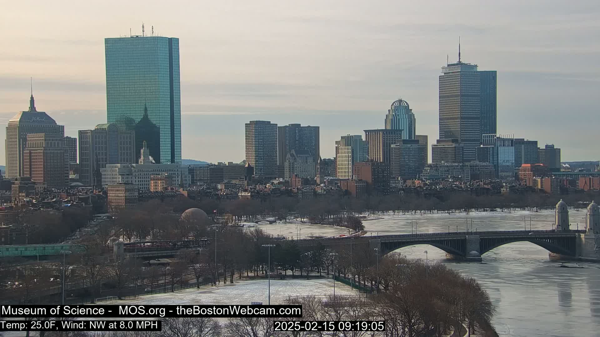 A partially frozen river flows beneath a bridge in front of a cityscape under a cloudy sky.