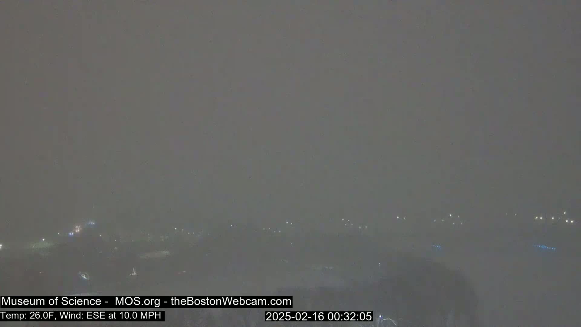 A hazy, snowy scene at night shows dimly lit distant buildings and grounds under a gray sky.