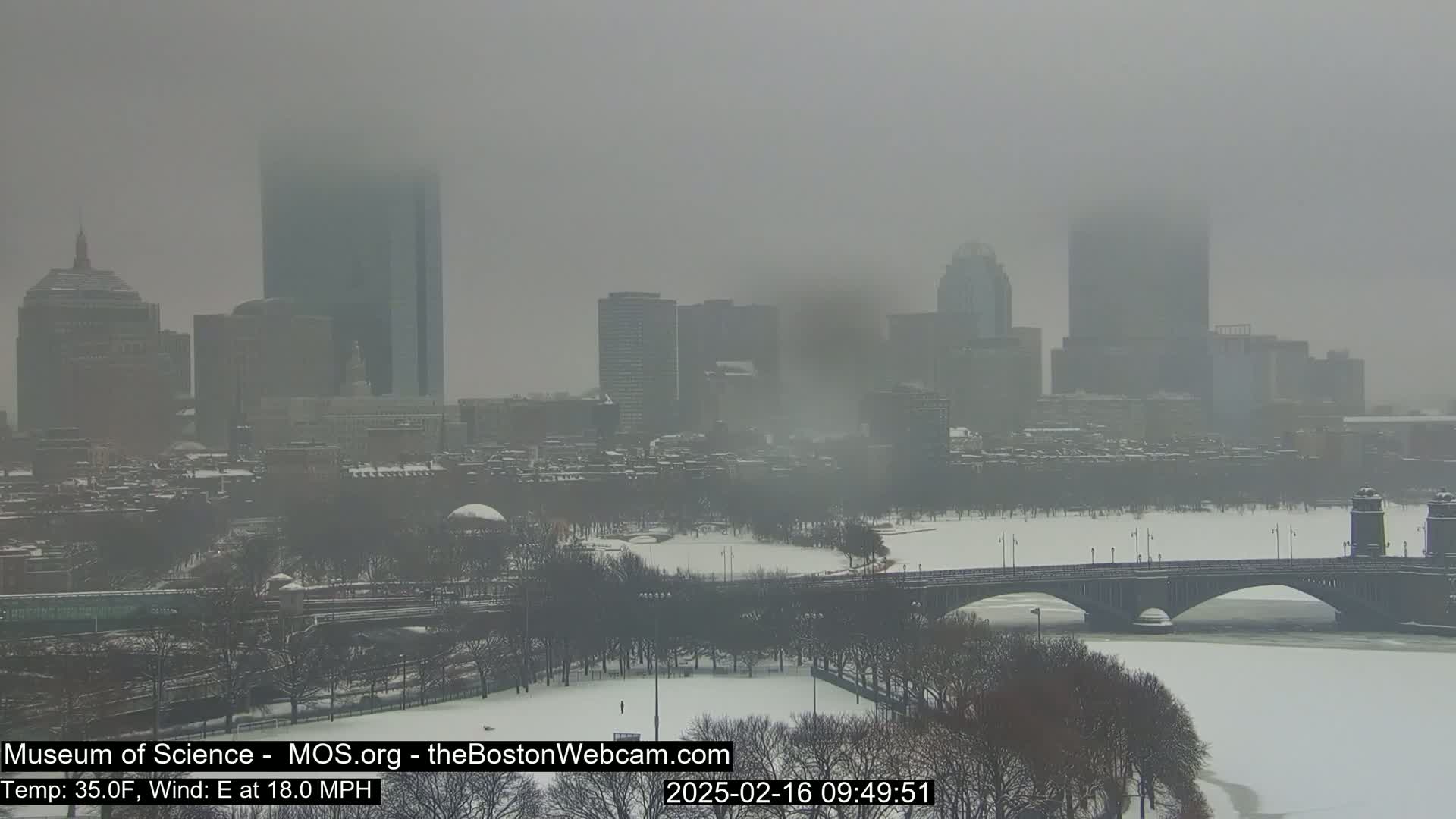 A snow-covered city skyline is partially obscured by fog, with a bridge spanning a frozen river in the foreground.