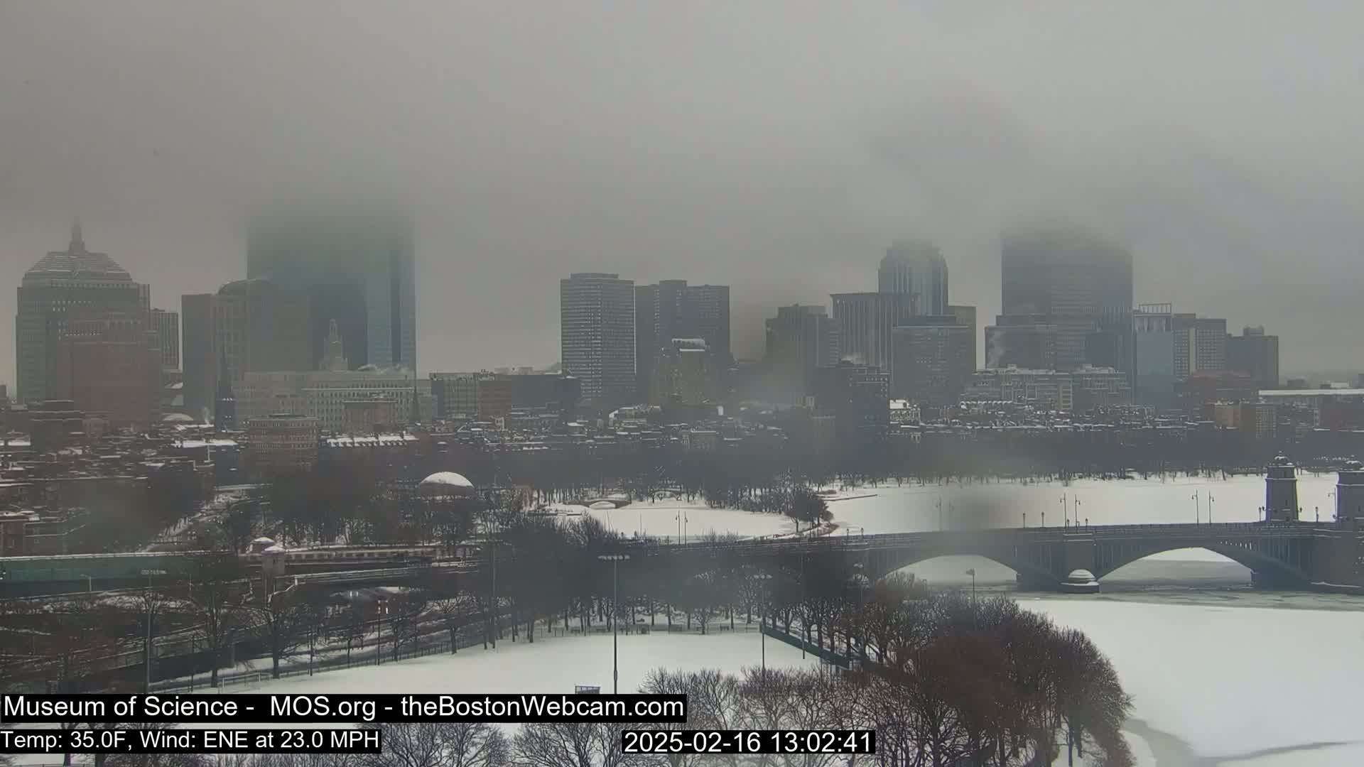 A snow-covered city skyline is seen through a hazy, foggy atmosphere, with a bridge spanning a frozen river in the foreground.