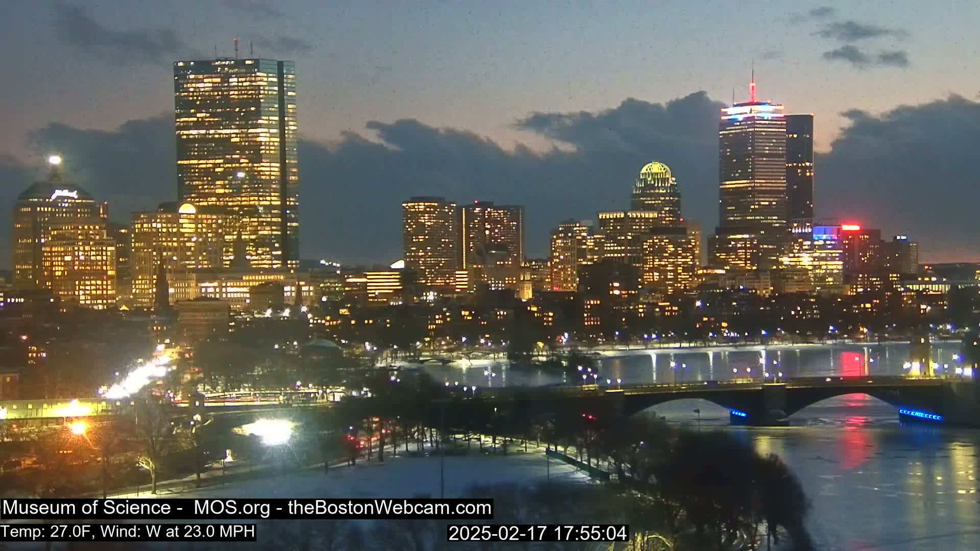 A nighttime cityscape with a river, bridge, and snow-covered ground under a partly cloudy sky, with a 27°F temperature and 23 mph westerly wind.