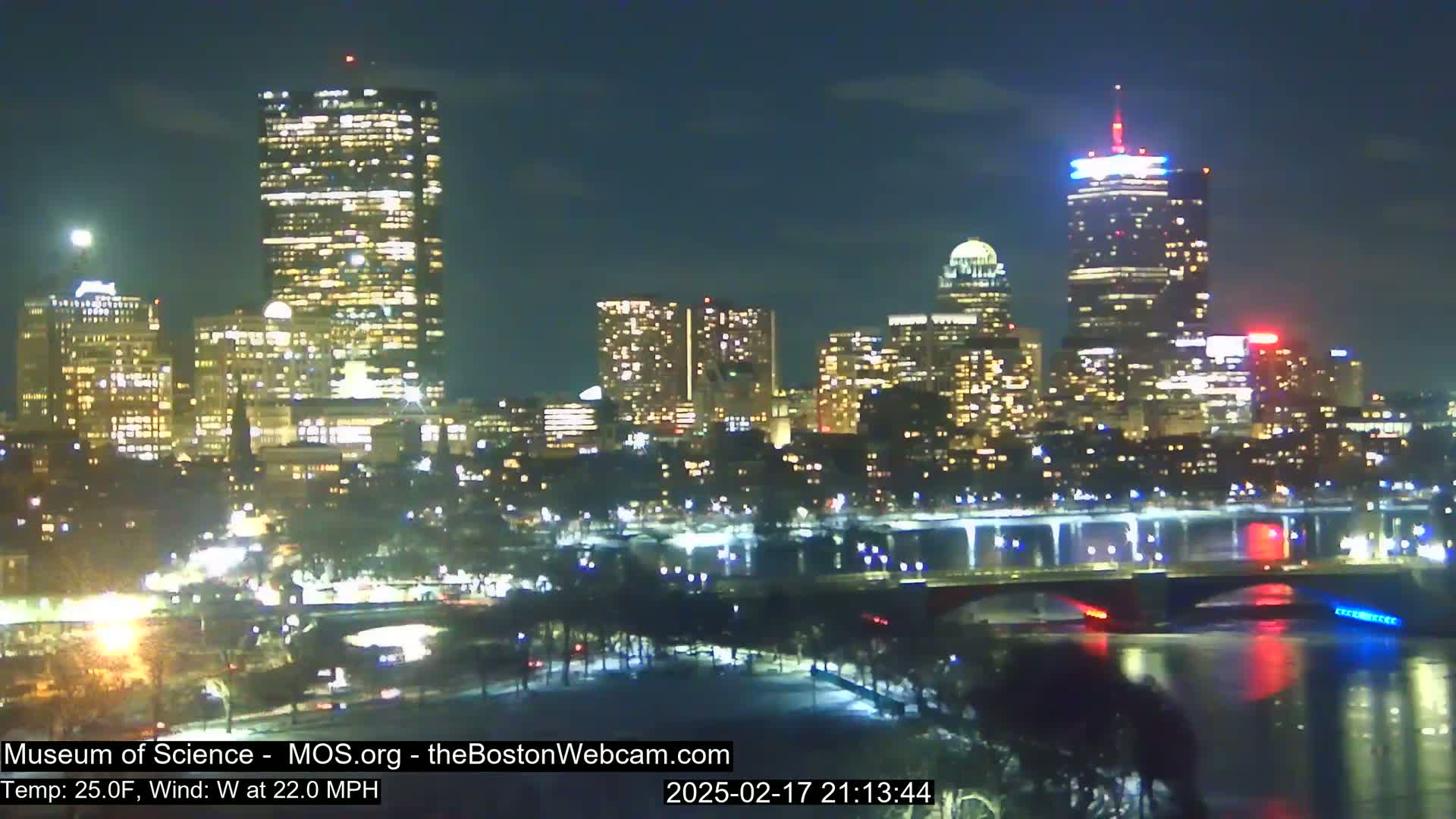 A nighttime cityscape with illuminated buildings and bridges reflected in a dark river under a clear sky, with a west wind at 22 mph and a temperature of 25°F.