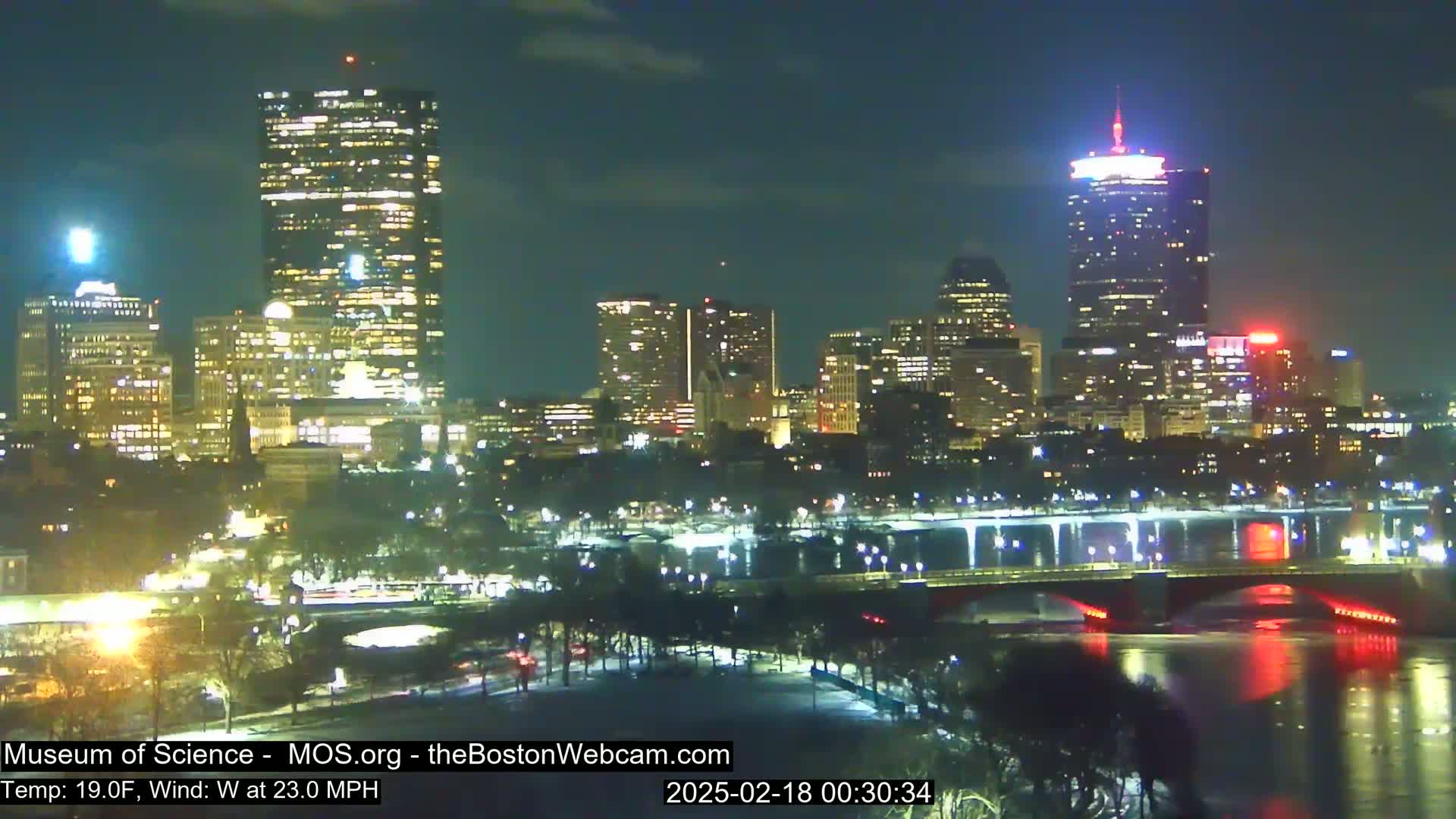A nighttime view of a city skyline with brightly lit skyscrapers across a river, under a clear sky and with a 19°F temperature and a 23 mph west wind.