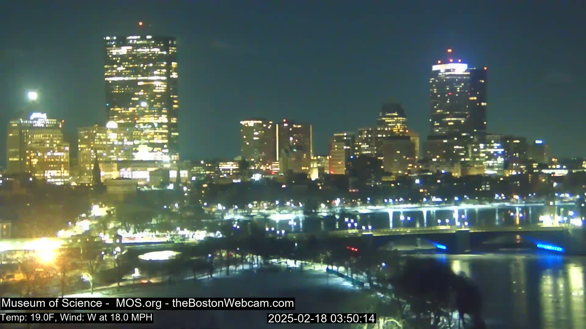 A nighttime view of a city skyline across a body of water under a clear sky with a 19°F temperature and a 18 mph west wind.