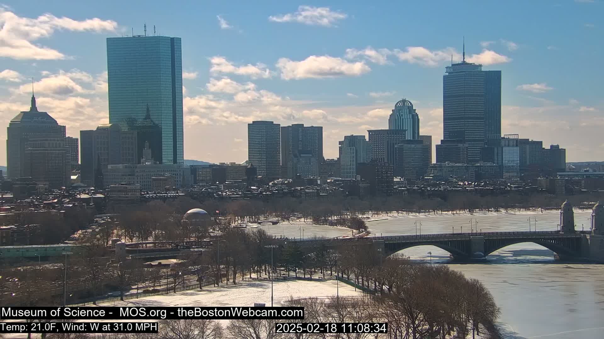 A partly cloudy, cold, and windy day shows a cityscape with several skyscrapers and a bridge spanning a partially frozen river, surrounded by snow-covered trees and parkland.