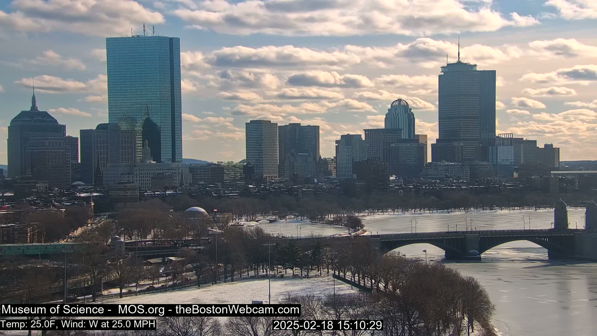 A partially frozen river flows beneath a bridge in front of a cityscape under a partly cloudy sky.